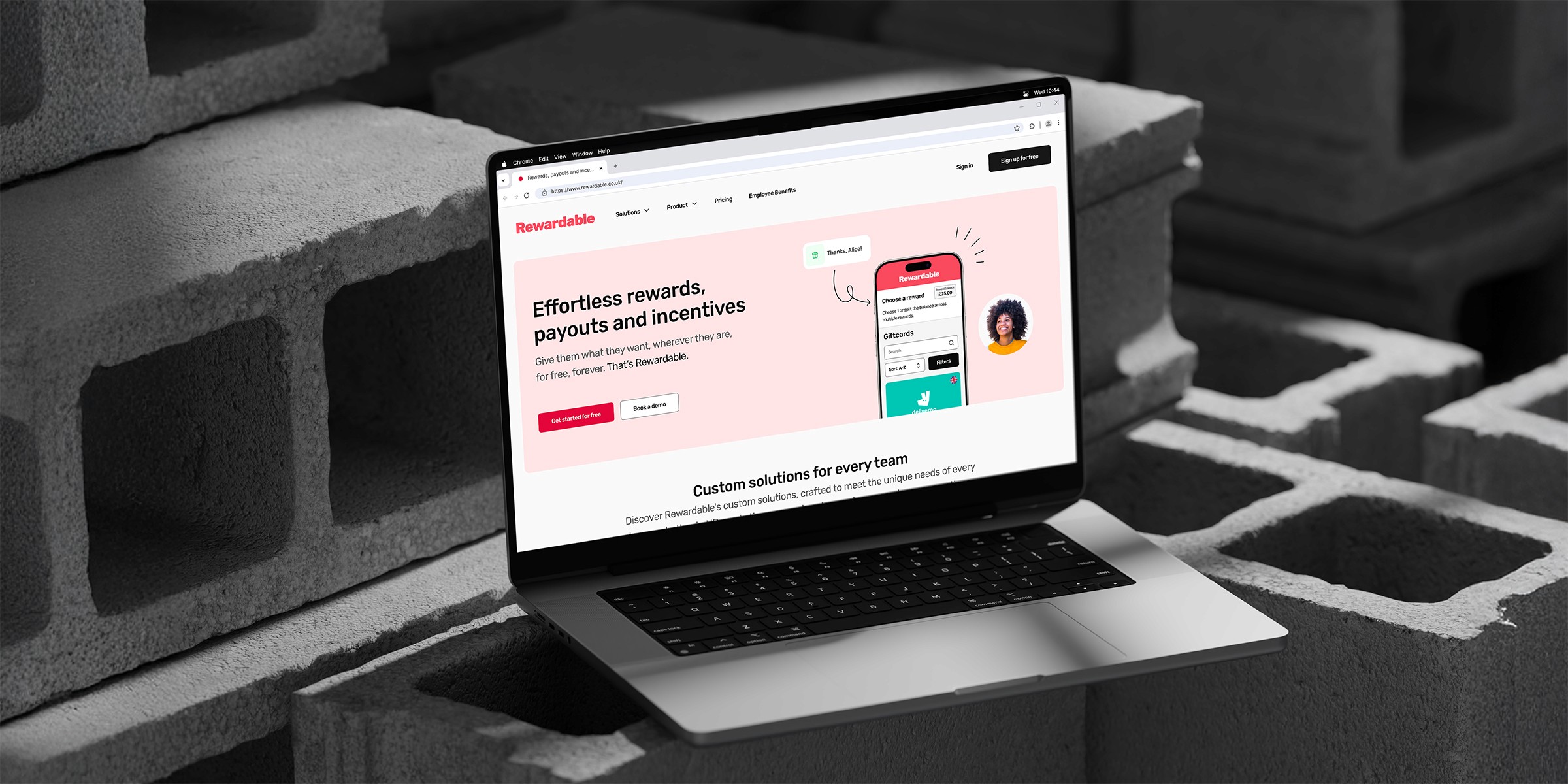 A sleek laptop displaying Rewardable's website about rewards, payouts, and incentives, set against a backdrop of concrete blocks, highlighting a digital e-commerce platform for gifting.
