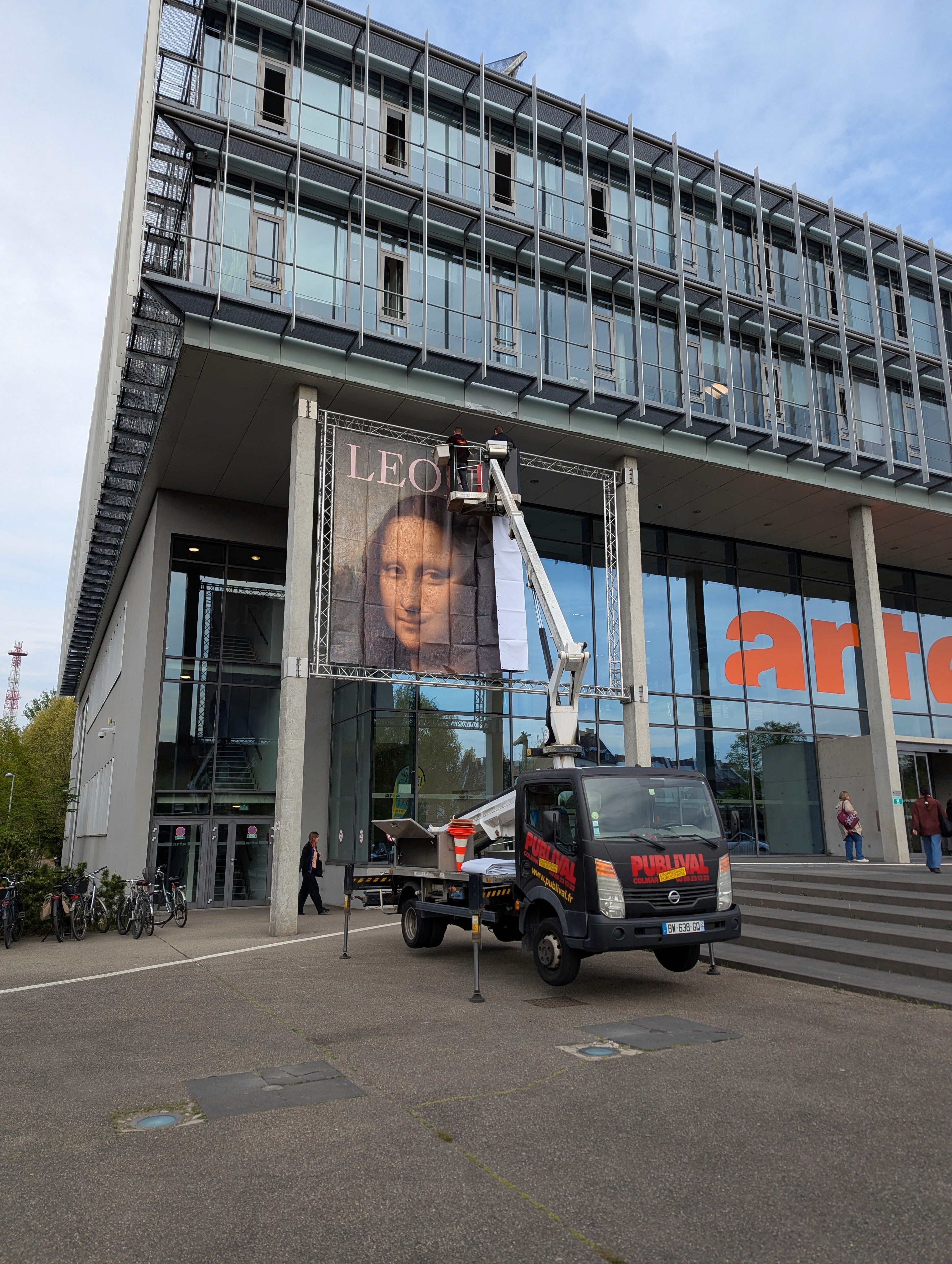 A large banner featuring a portrait of Mona Lisa is being hung on a modern building with 'arte' written on it by workers in an aerial lift.