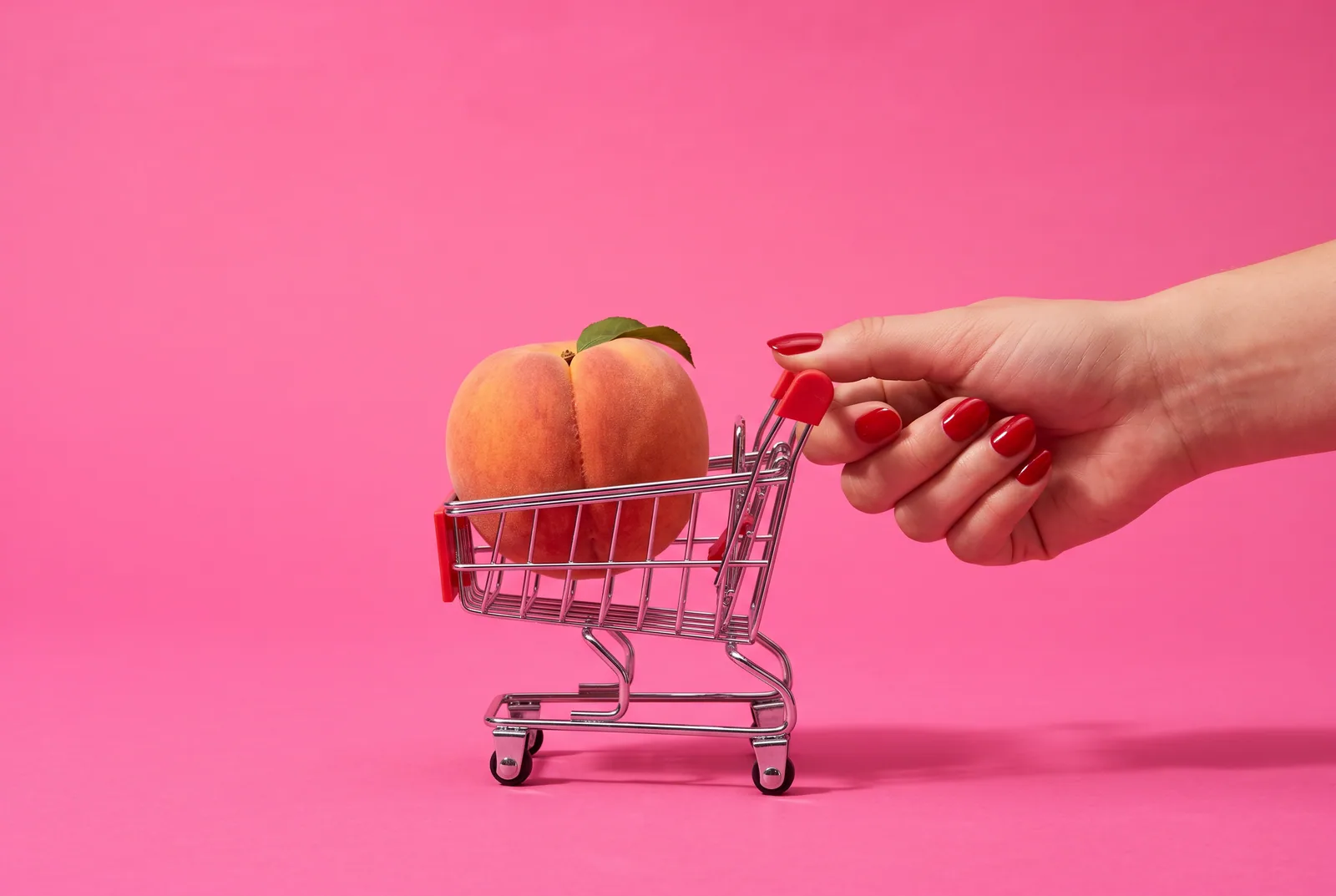 OnlyFans addiction concept: a ripe peach sitting inside a tiny chrome shopping cart on a hot pink backdrop