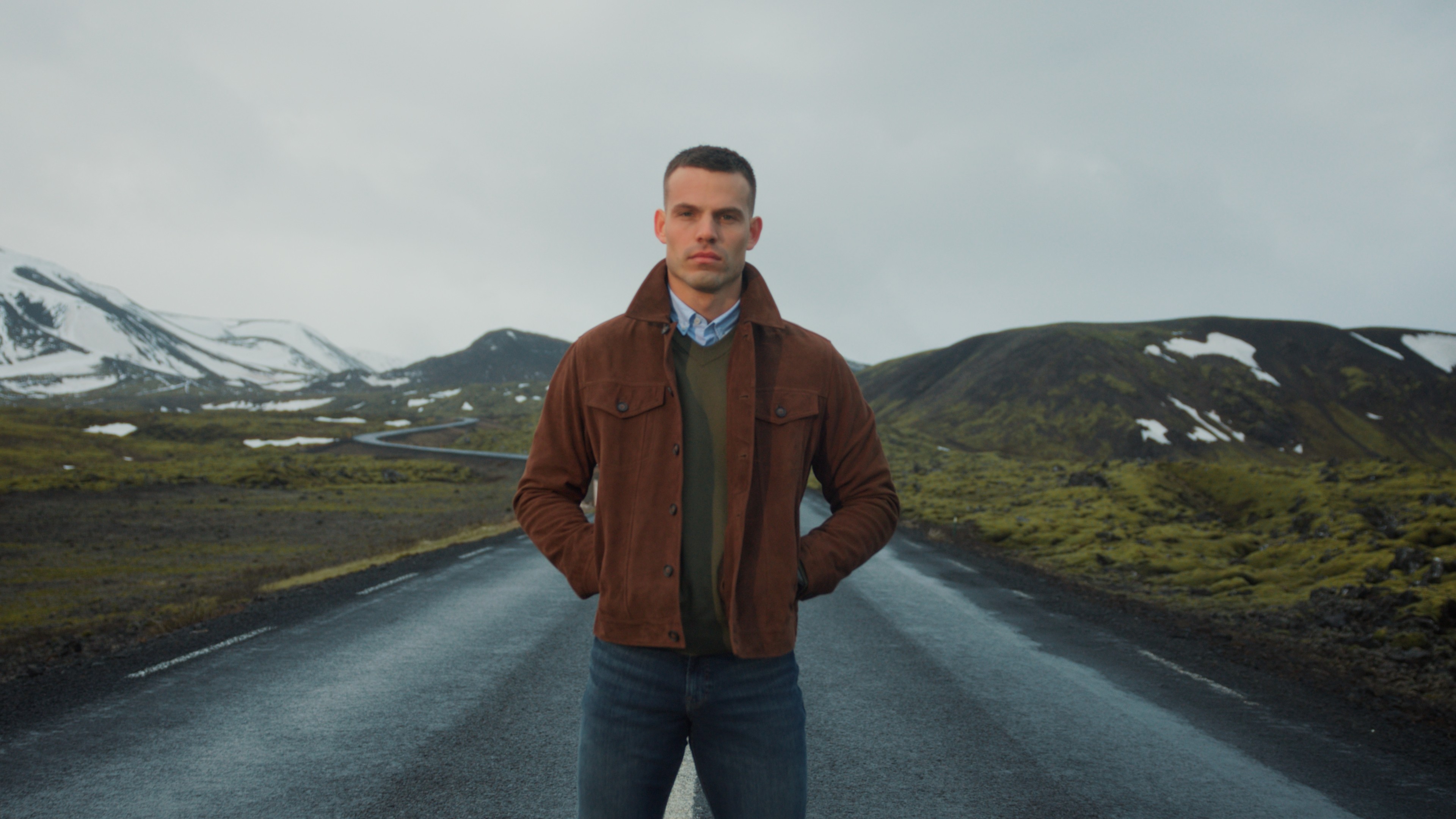 Man in a brown suede jacket standing on an empty road below snow-capped peaks