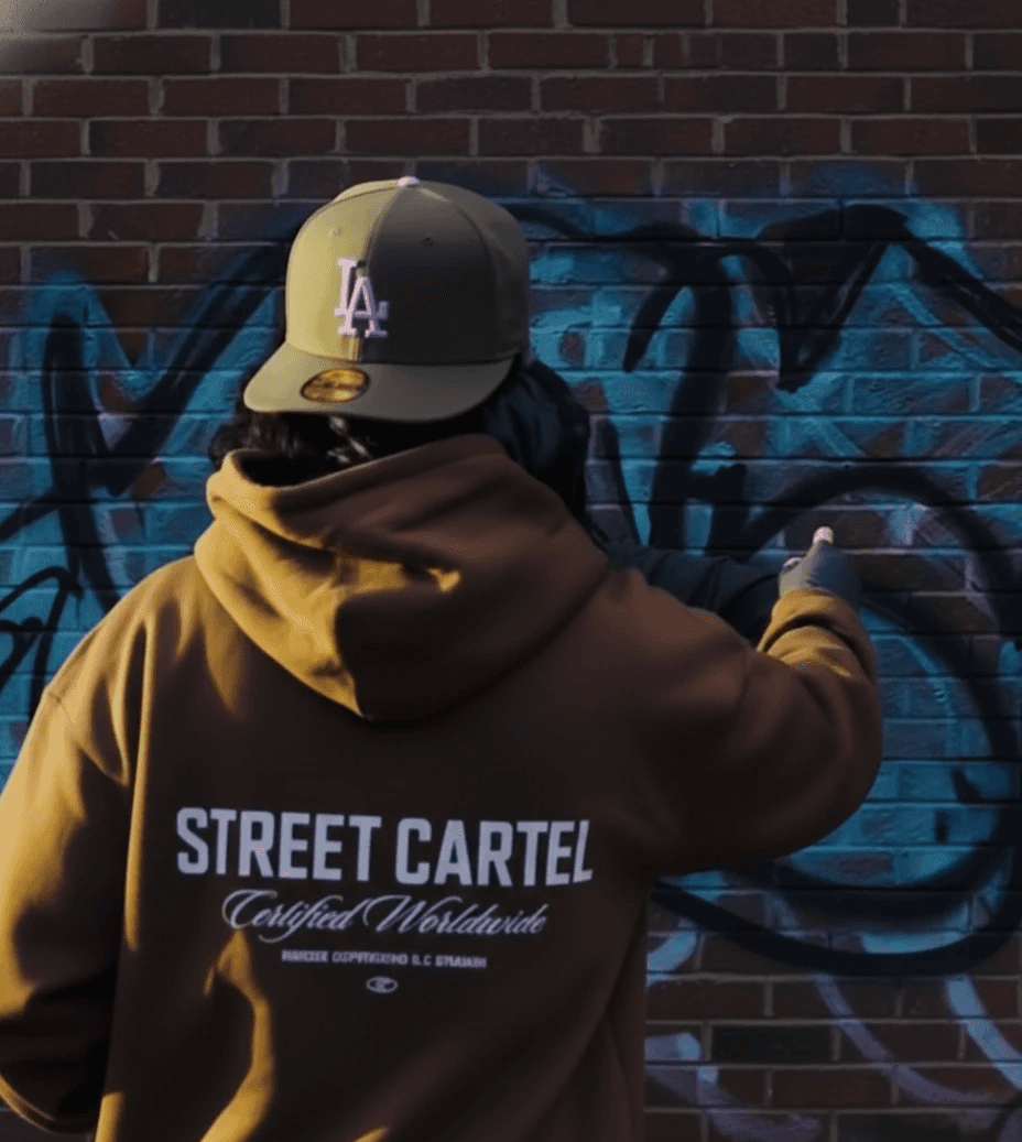 Streetwear photo of a model wearing a mustard Street Cartel hoodie against a graffiti wall.