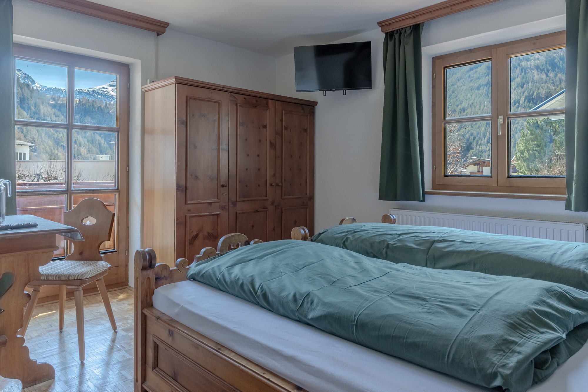 Ein gemütliches, rustikales Schlafzimmer verfügt über ein Doppelbett aus Holz mit grüner Bettwäsche, einen passenden Kleiderschrank, große Fenster mit Bergblick und einen an der Wand montierten Flachbildfernseher.