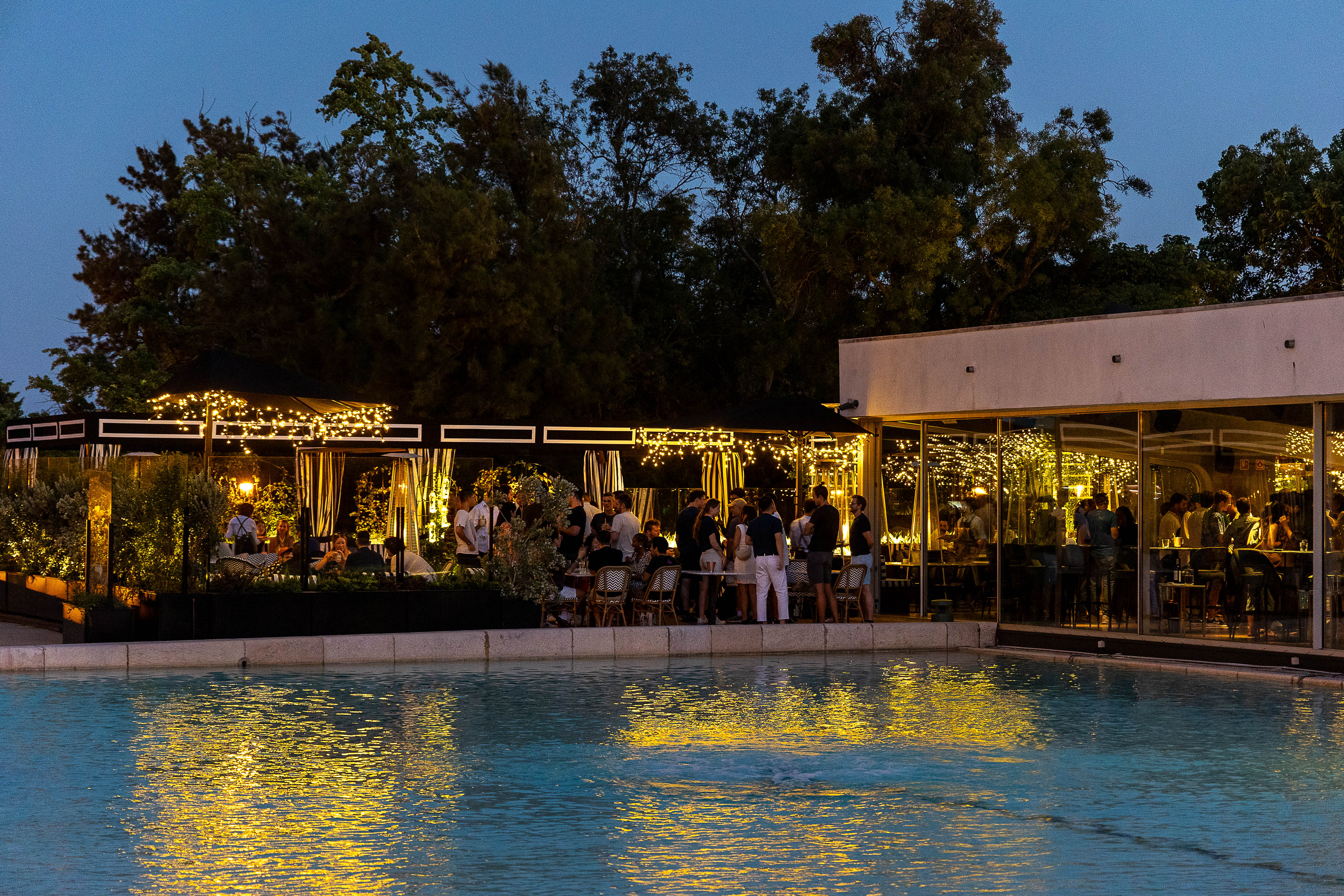 an outdoor evening scene at a venue by a pool. Soft lighting from the trees and patio areas creates a warm and inviting atmosphere. People are gathered around tables, indicating a social event or gathering.