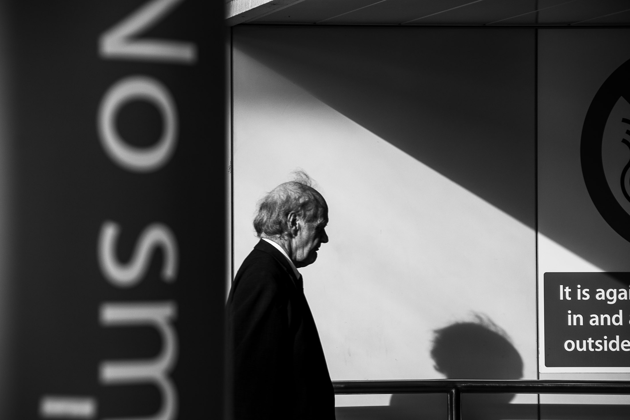 Black and white photograph of an elderly man walking past a wall with strong geometric shadows.