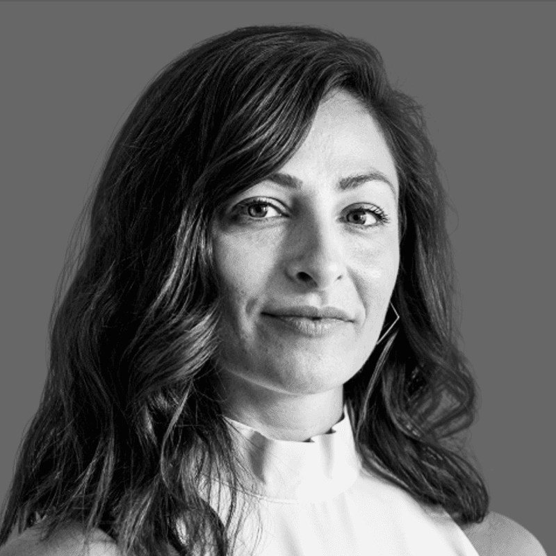 A man reading from a tablet in a collaborative office environment. Desks with documents and a discussion between colleagues are visible in the background.