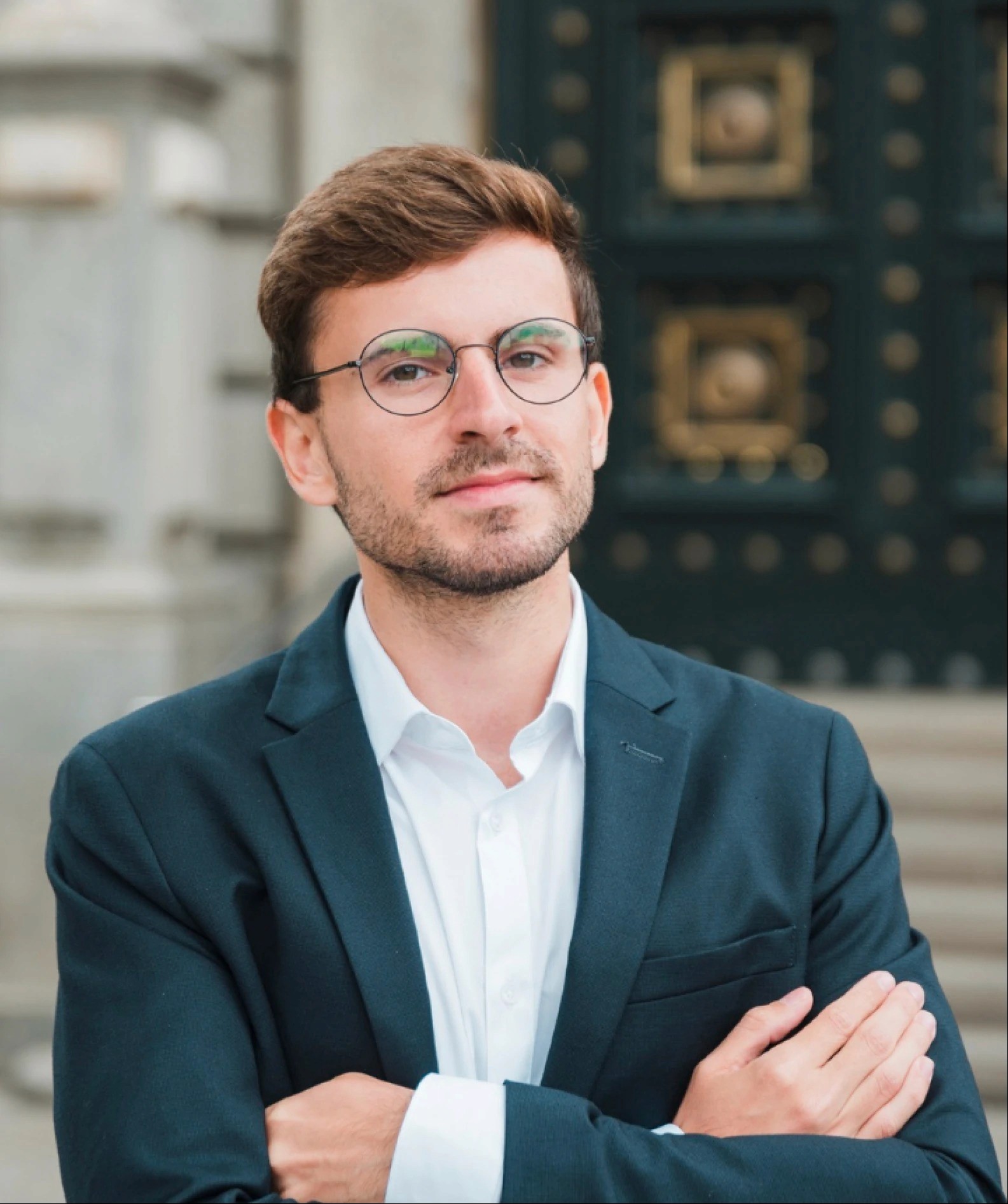 A young man with glasses and light brown hair stands confidently, arms crossed, in front of a decorative building.