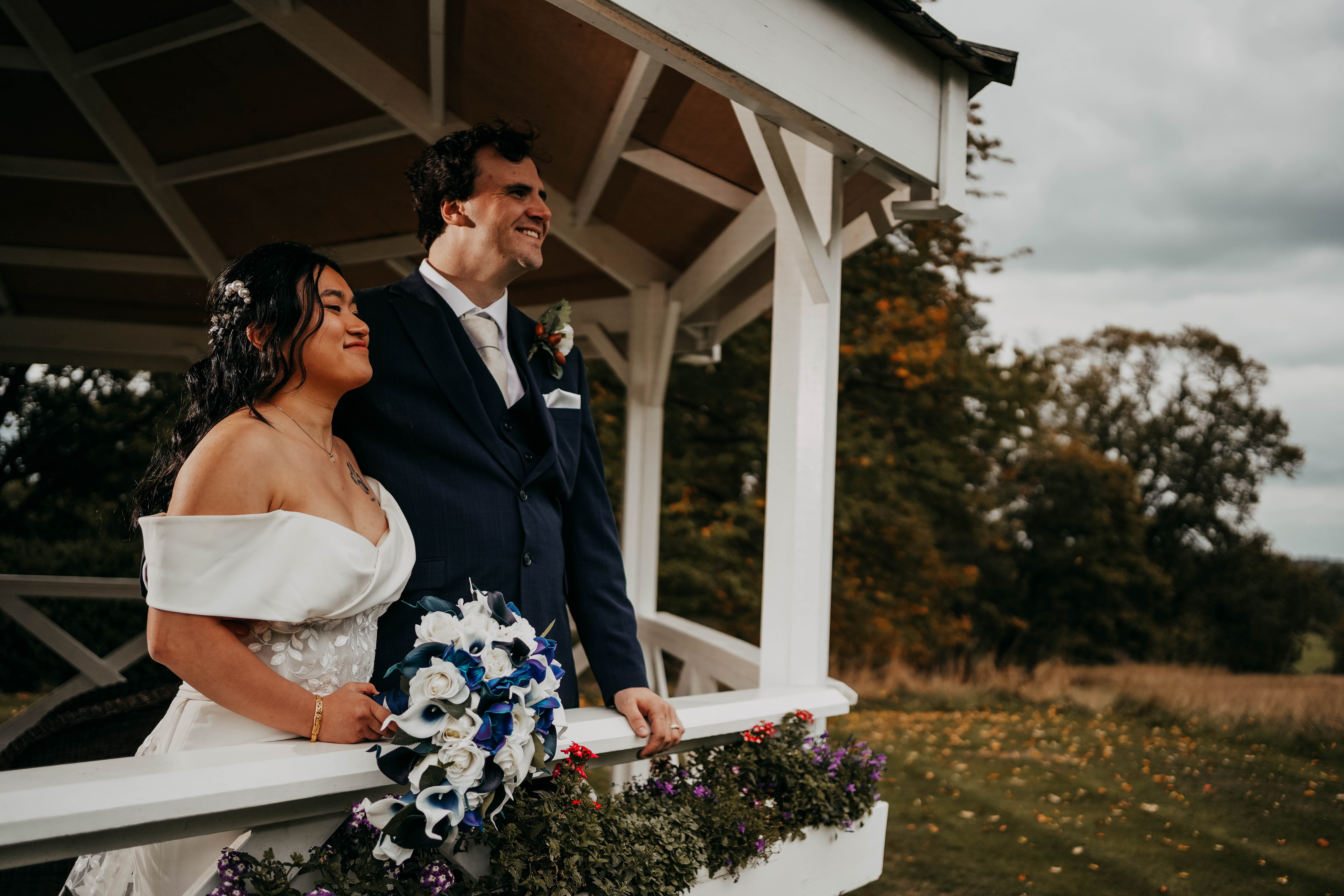 The bride and groom stand together in a white gazebo, smiling as they look out at a field with trees in the background. The bride holds her blue and white bouquet as she leans on the railing.