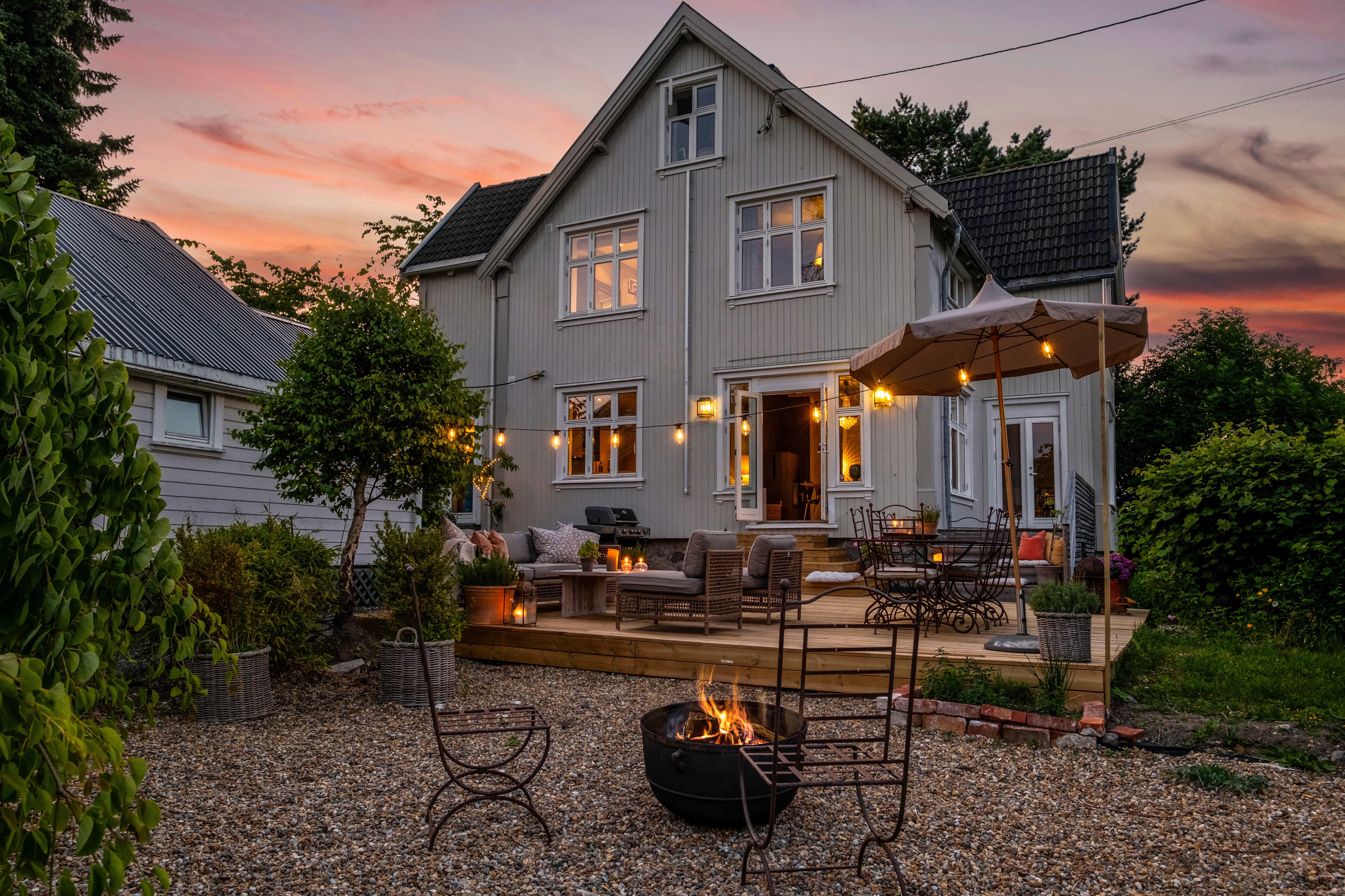 Hus med treterrasse, utemøbler og bålpanne i hagen ved solnedgang
