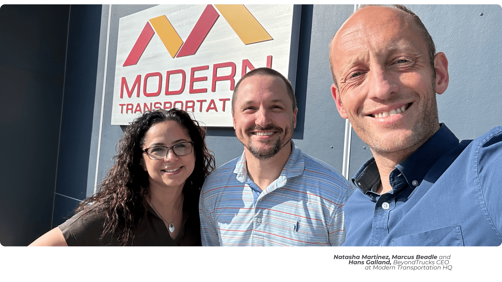  three people smiling in front of the entrance of a company. From left to right, a woman with curly hair, glasses and a black shirt, in the center a man with a beard and short hair, wearing a light-colored dress shirt, on the far right, a bald man, wearing a darker blue dress shirt. In the background you can read in large letters what would be the name of the company "Modern Transportation