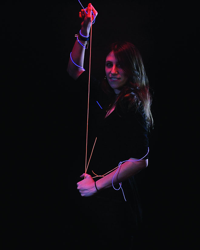 A woman with long brown hair holds a thin, light-colored string or wire in her right hand. She wears a dark long-sleeved top and several bracelets on her left wrist. The background is entirely black, and the string is illuminated with red and blue lights along its length. The woman’s face is partially visible, and she appears to be smiling.