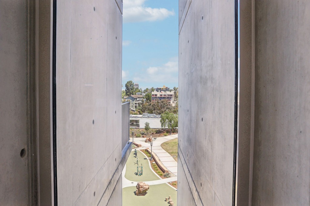 View through narrow concrete walls reveals Olive Street Park with green pathways, sculptures, and distant buildings under a sky with scattered clouds. Calm and modern.