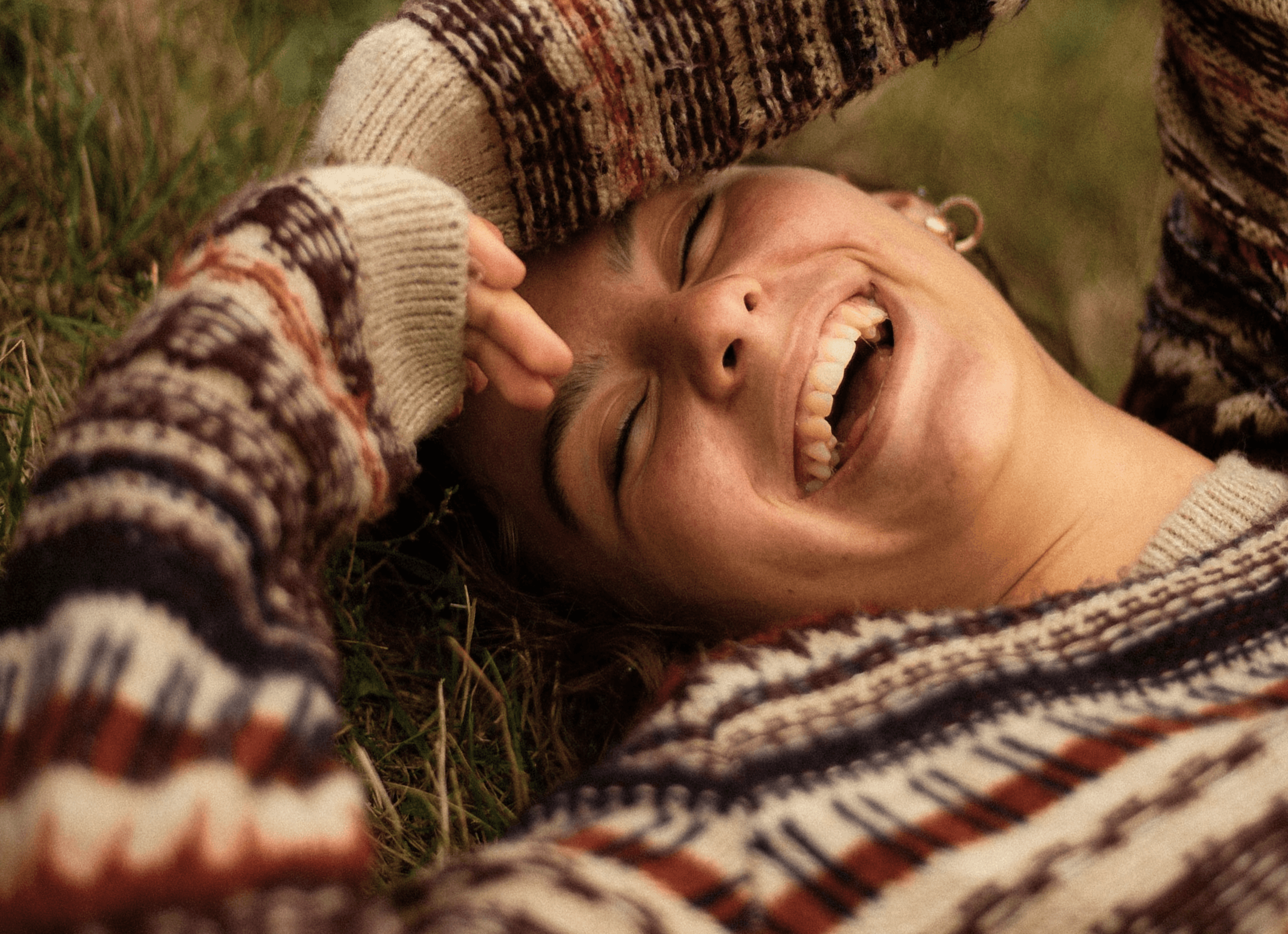 smiling woman in brown and white sweater lying on green grass field during daytime