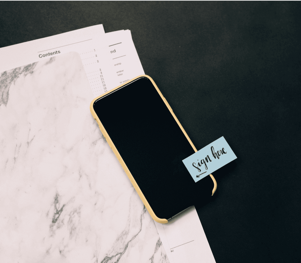Ein Handy mit einem Sign here Post-It liegt auf einem Tisch über Verträgen