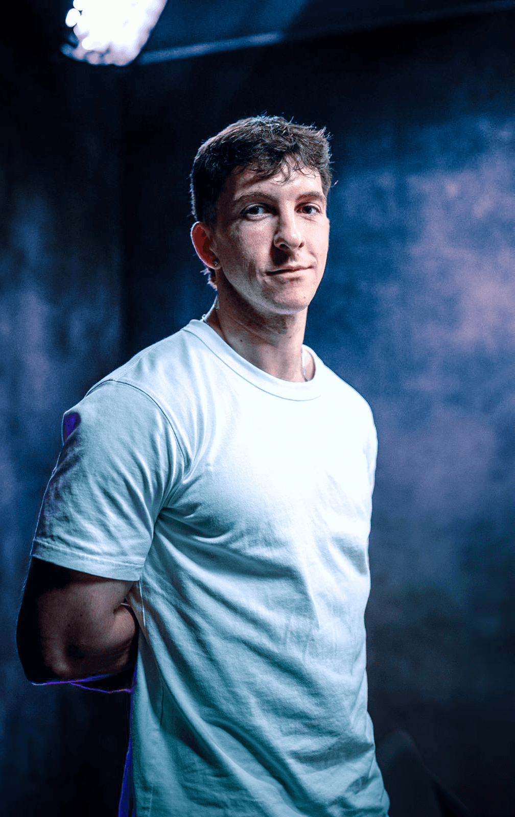 Portrait professionnel et moderne d'un jeune homme, représentant le fondateur ou directeur artistique de Nux-Studio, posant de face avec un léger sourire. Il porte un simple t-shirt blanc à manches courtes. L'arrière-plan est un mur texturé, sombre et baigné d'un éclairage dramatique de couleur bleu électrique et violet, créant une ambiance de studio créatif et innovante. L'espace négatif important sur la gauche est idéal pour l'ajout d'un titre, d'un logo, ou d'un slogan. Ce visuel évoque l'expertise en branding, direction artistique et l'identité moderne d'une agence de design digital.