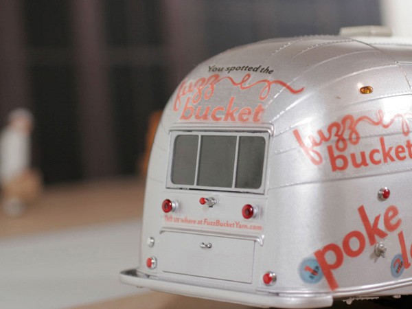 A close-up of a silver model bus with “fuzz bucket” painted on its side in red cursive script. The bus has three red lights on the back and a small window on the side. A blurred figure is visible in the background to the left. The bus sits on a light-colored surface.
