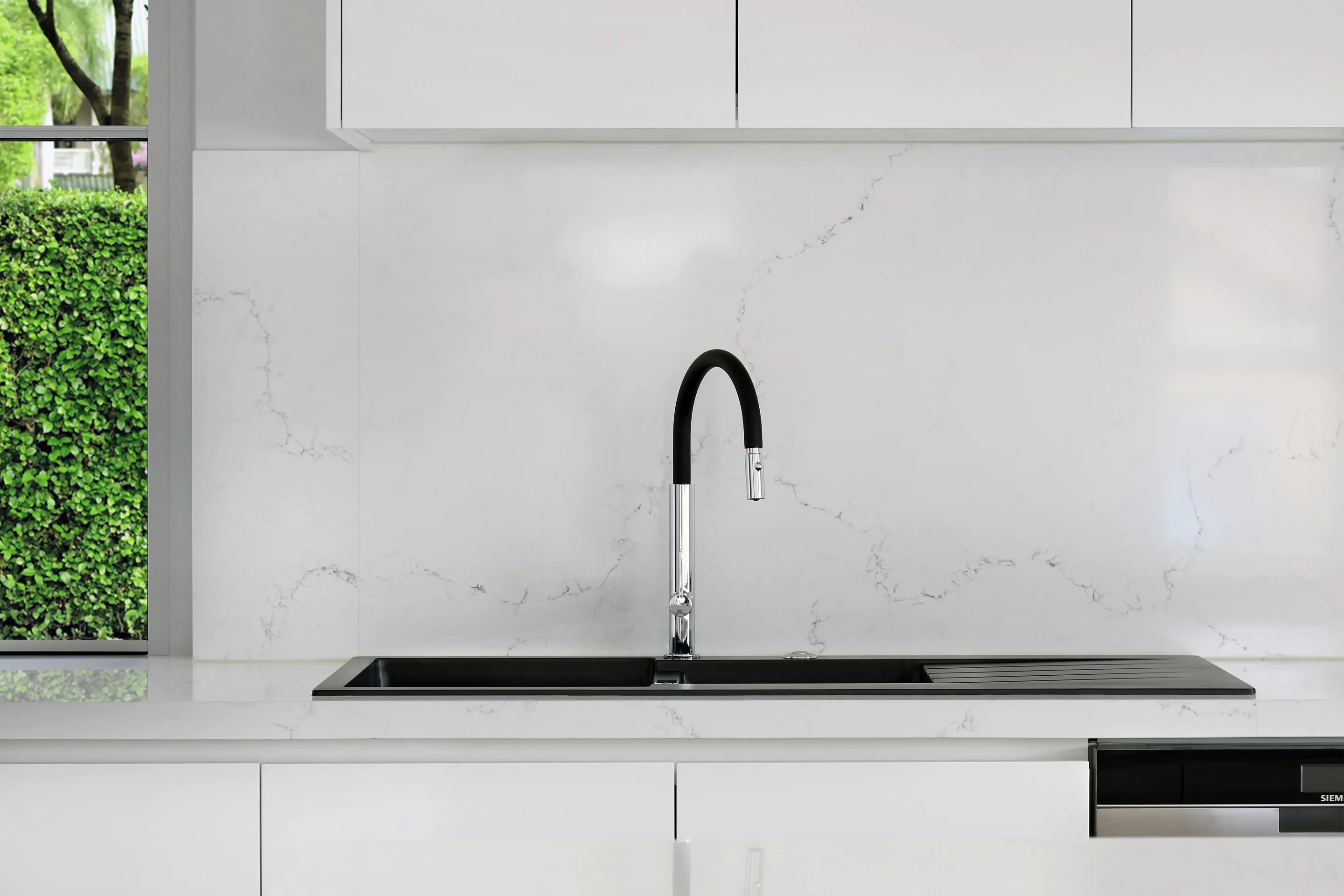 Kitchen benchtop detail with a white veined stone splashback, black undermount sink and drainer and a black-and-chrome gooseneck tap by a window onto a hedge