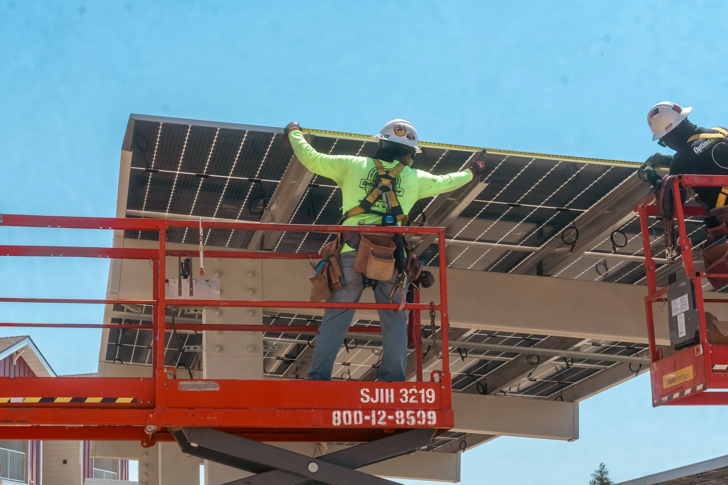 Two installers on a lift guiding a solar panel into a carport frame against a blue sky