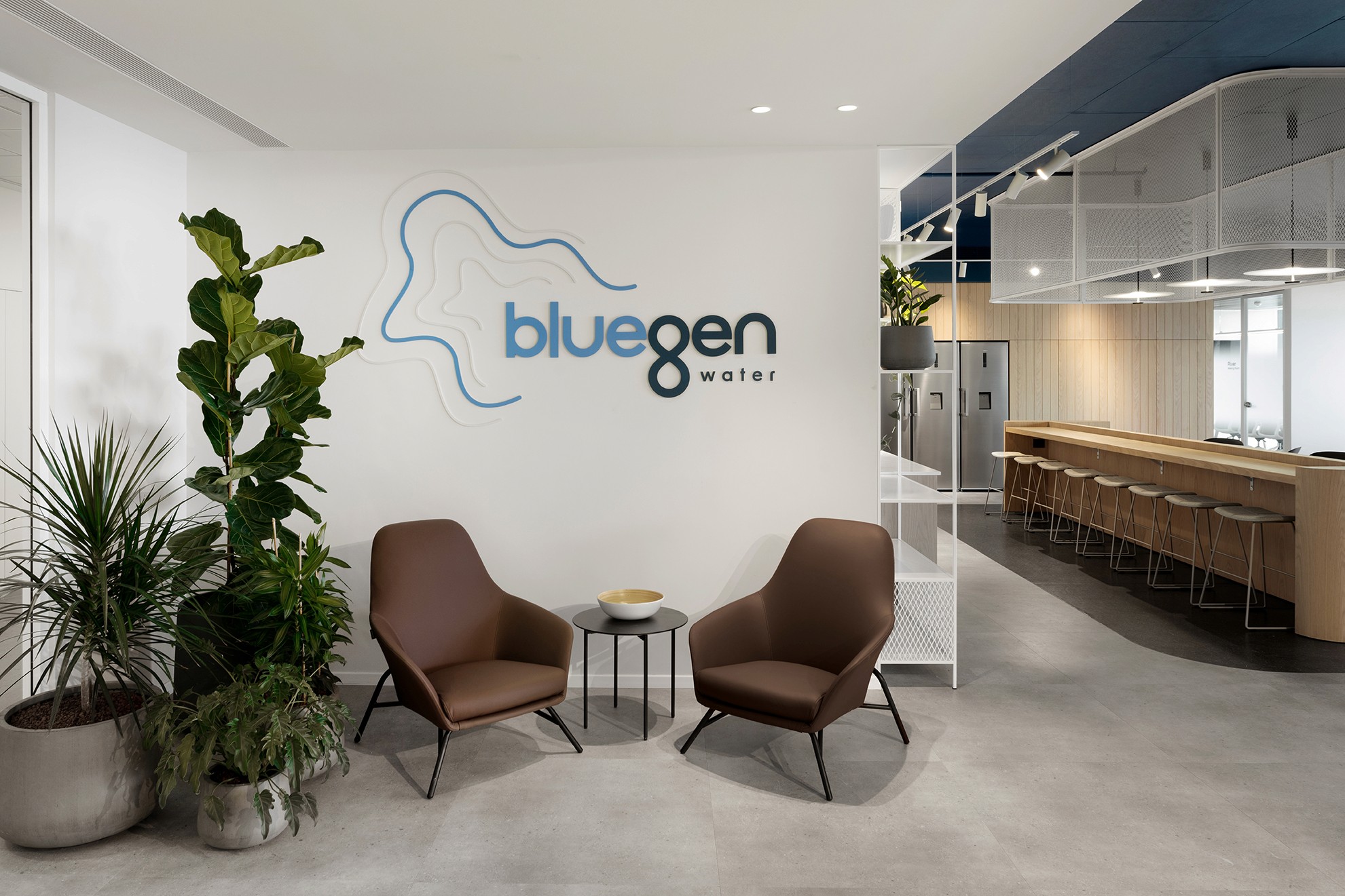 כניסה למשרדים: קיר מיתוג בייצור CNC תלת-ממדי בשילוב ריהוט רך (Soft Seating) ורמיזה לחלל המטבח התעשייתי מאחור