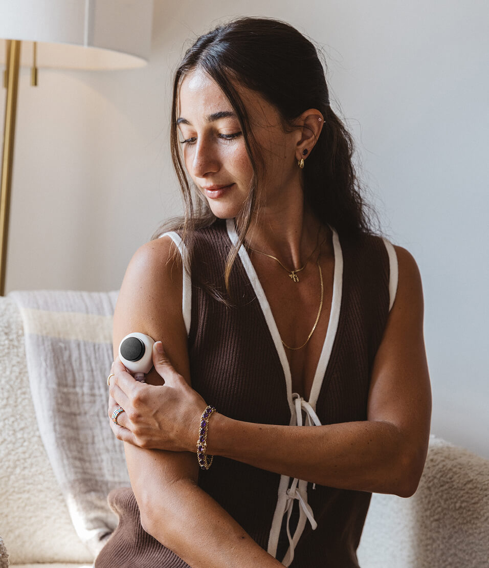 Woman using a Rythm at-home blood collection device on her upper arm, seated indoors in soft natural light, looking down as she applies the device.