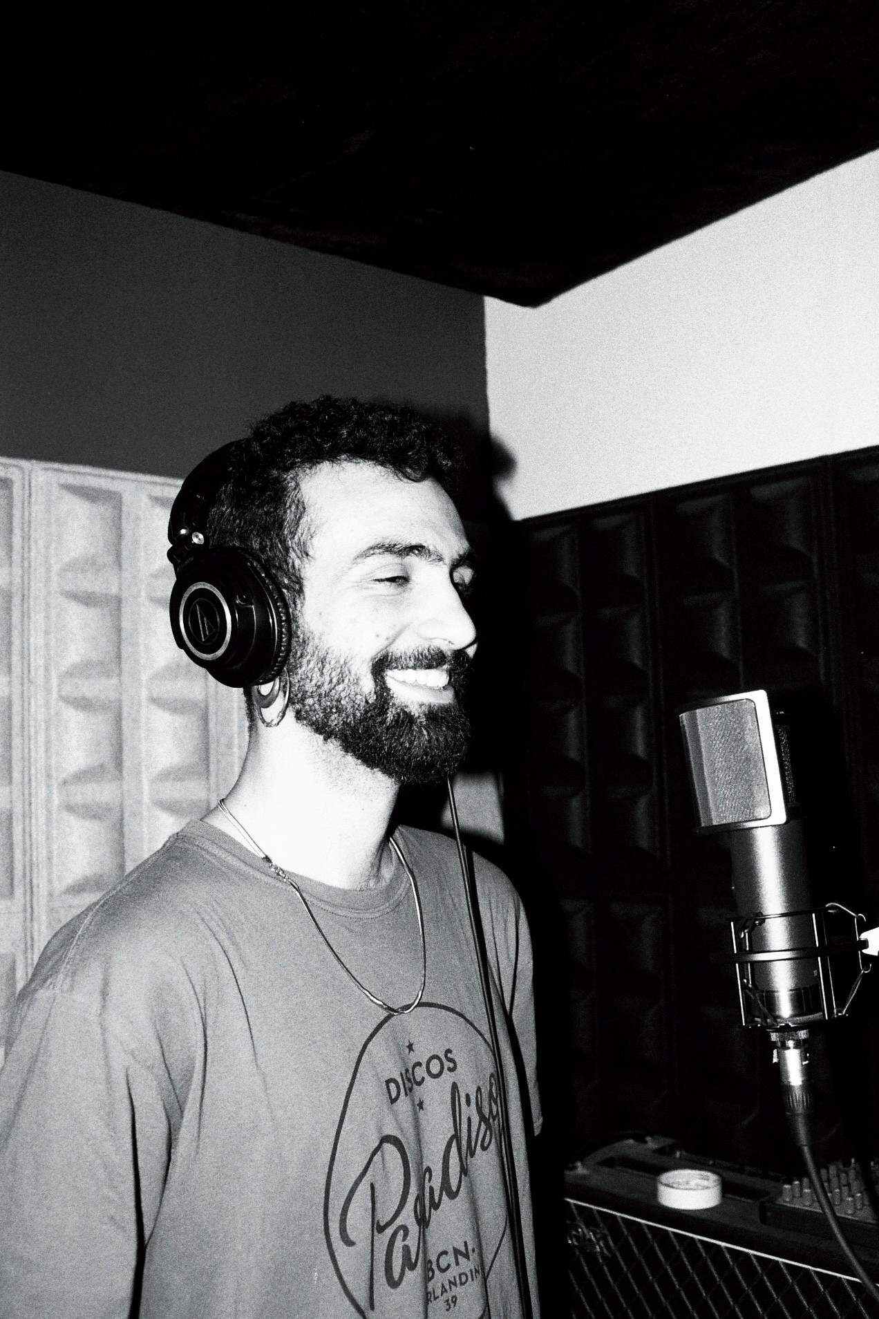 A smiling person with a beard and professional headphones stands in a recording studio, surrounded by soundproofing panels.