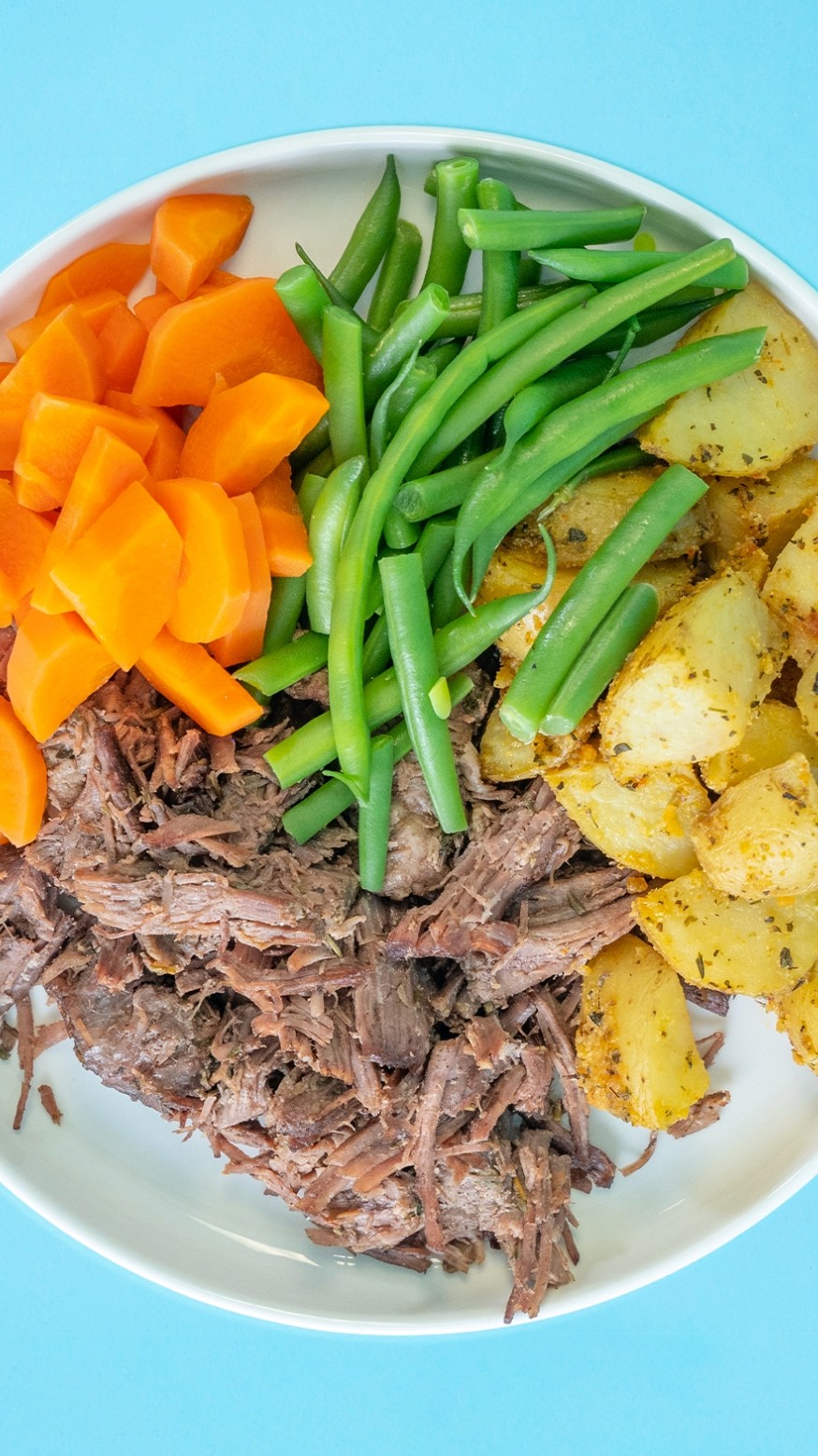 Plate of shredded beef roasted potato, green beans and carrots on a blue background