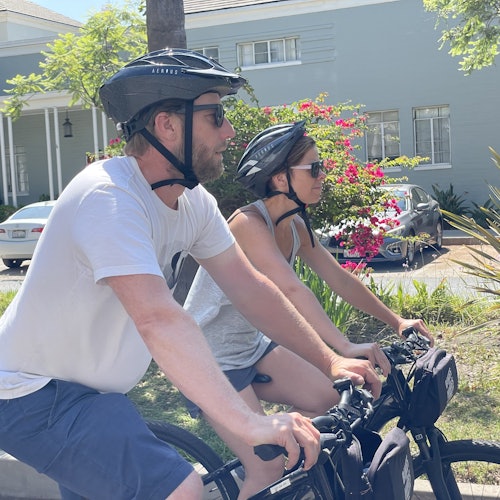 Two people wearing helmets and sunglasses ride bicycles on a sunny street, with a house and flowering bushes in the background.