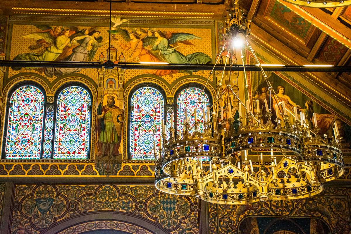 An ornate gold and blue interior of a church featuring stained glass windows, religious murals, and a large chandelier.