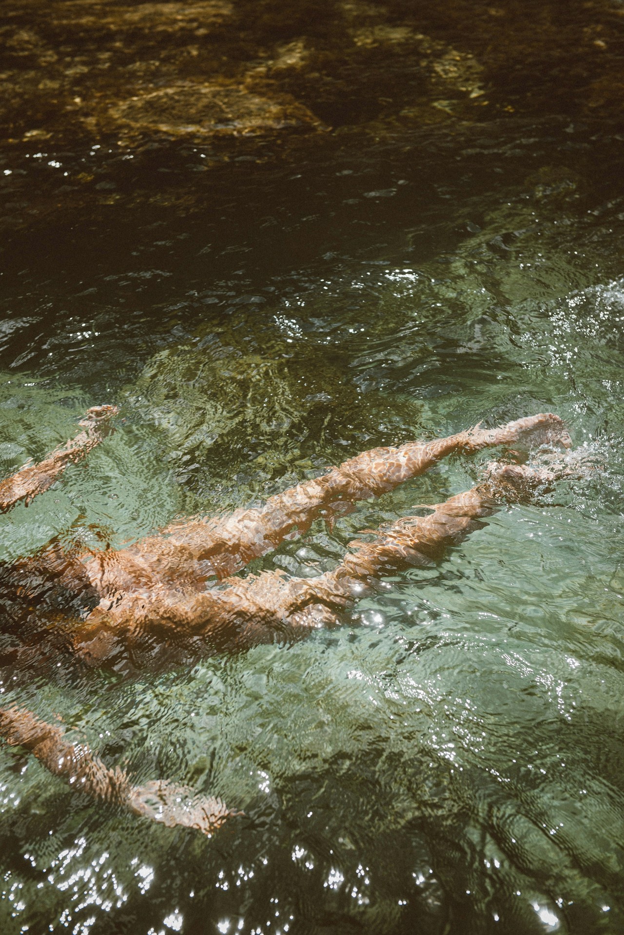 Jambes et bras d'une personne nageant dans une eau verte claire et ondulante. Des reflets de soleil scintillent.
