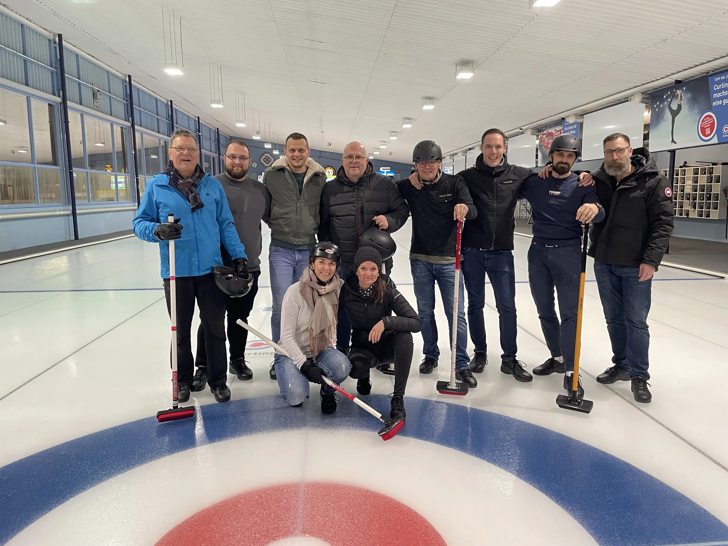 Team mit Curlingbesen auf dem Eis, im Vordergrund das rot-blaue Curling-Haus