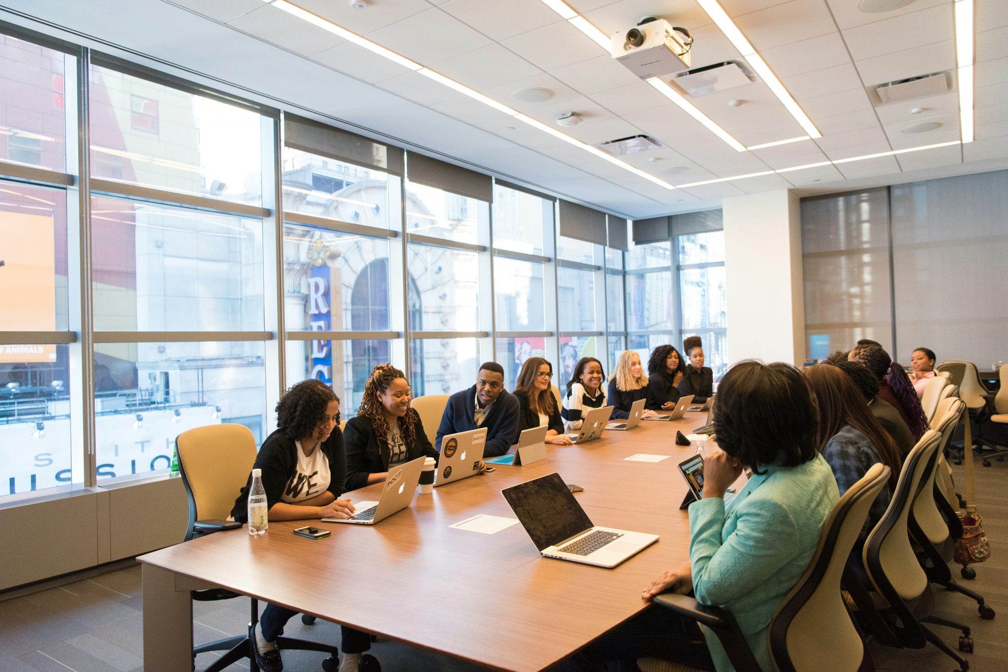 A diverse group of people seated at a conference table in a bright meeting room with large windows.