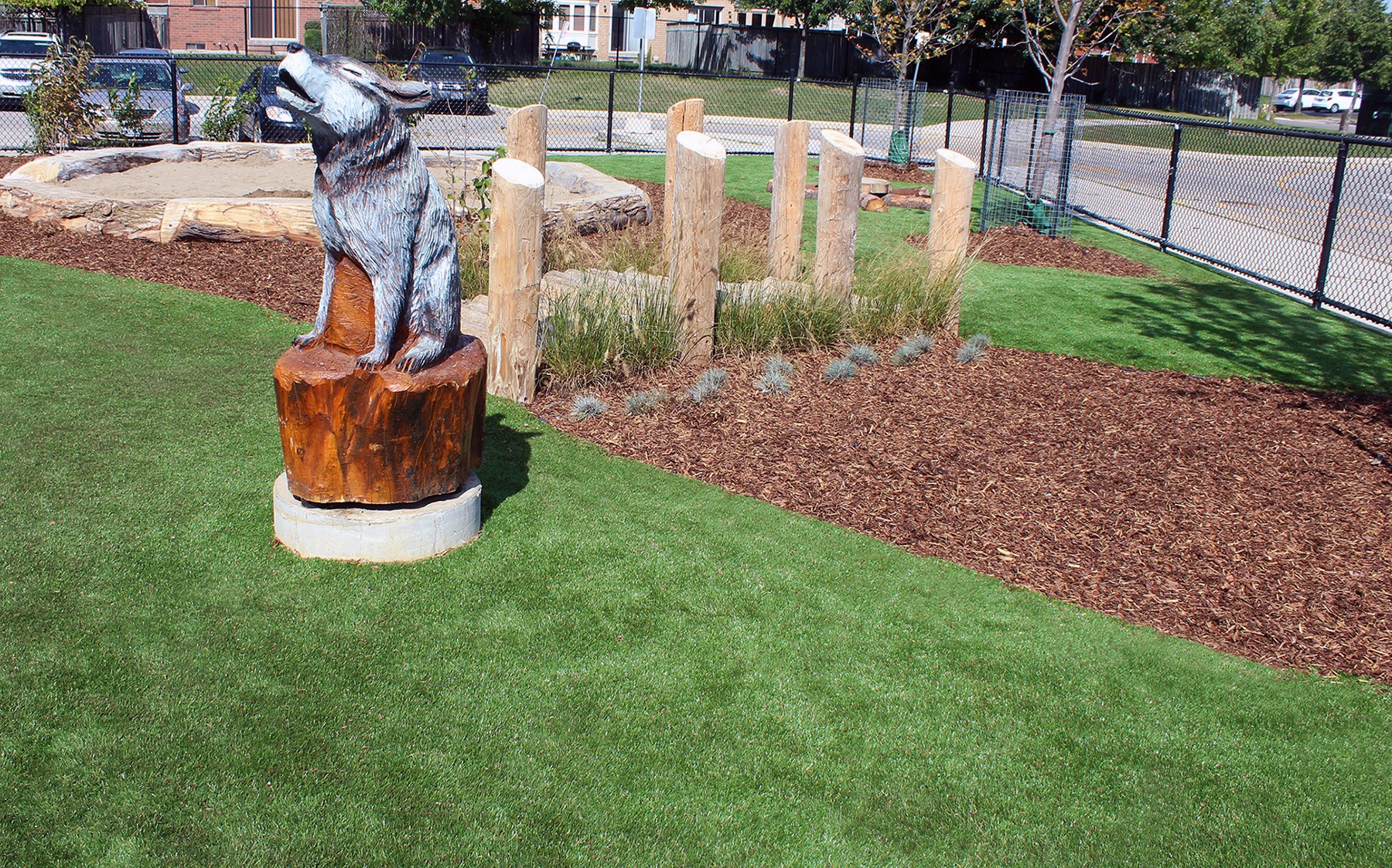 A dog statue perched on a wooden stump, surrounded by artificial AGL Grass in a peaceful playground setting.