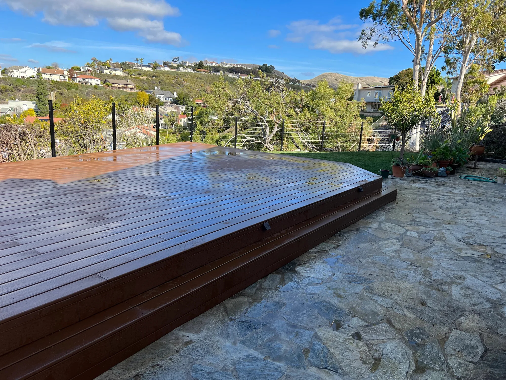 Two surfaces, one setting Timber deck beside an irregular stone-paved terrace with cable railings and a hillside outlook.