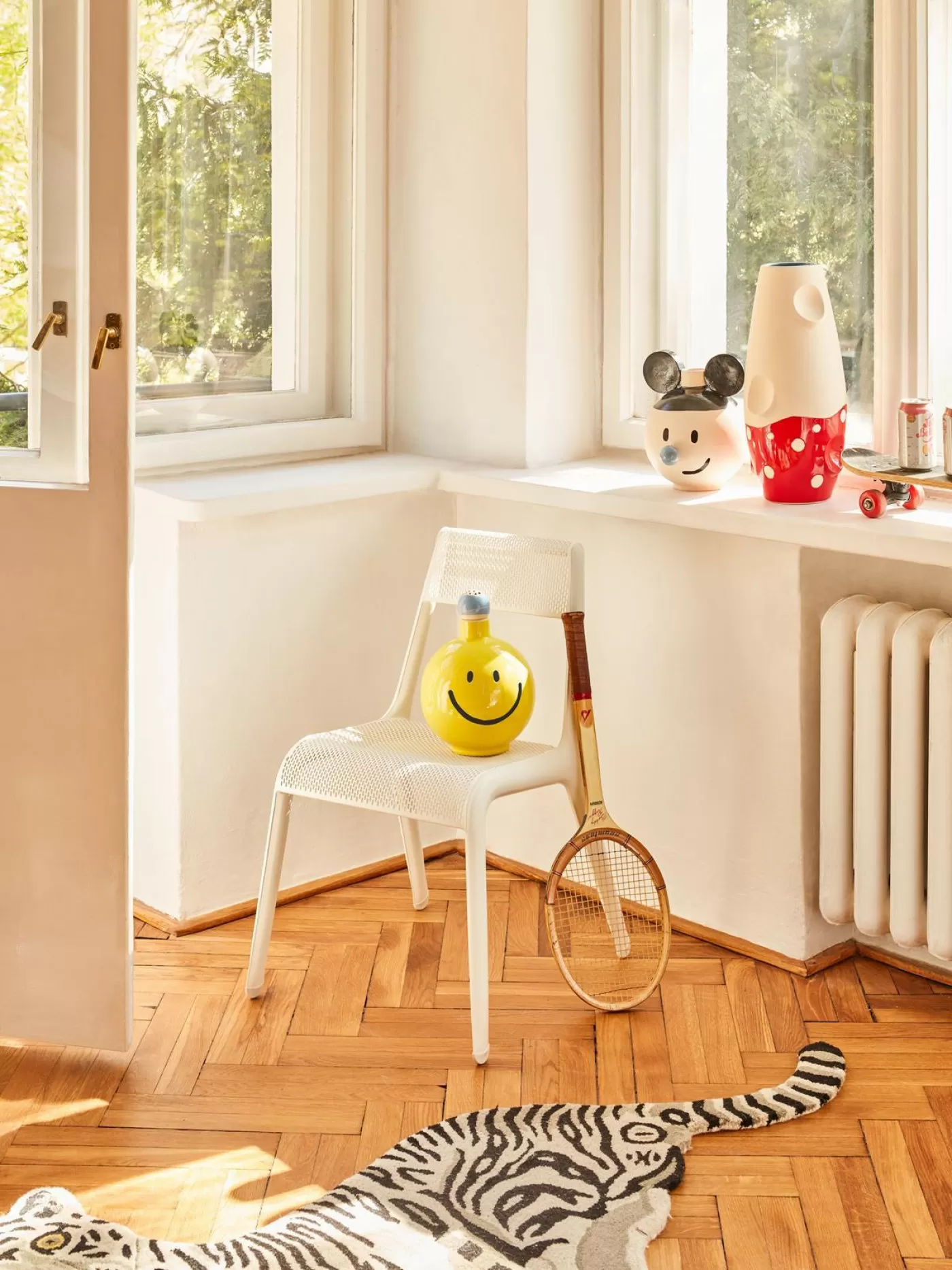 Bright room with large windows, a small chair, a yellow vase, and a colorful rug on wooden flooring.
