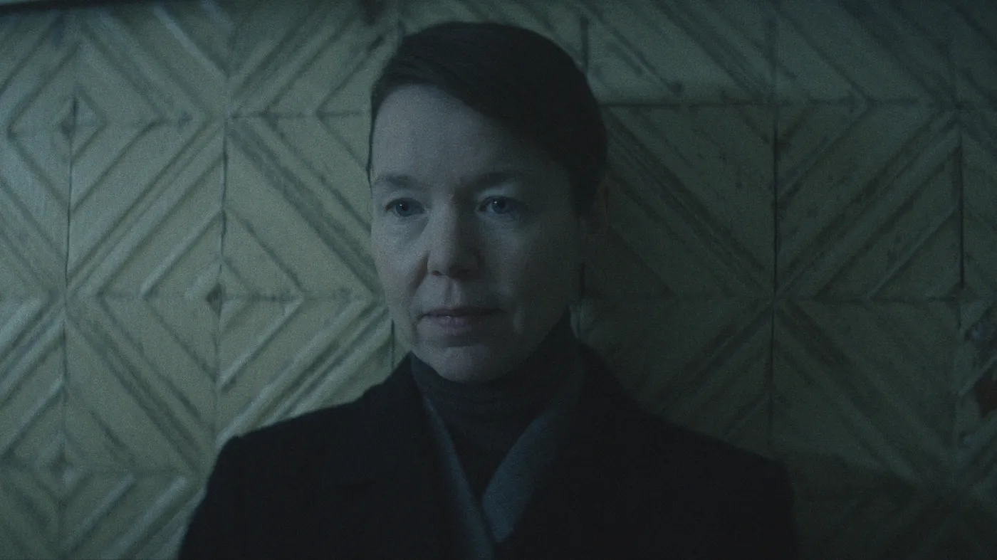 A woman with short hair and a neutral expression is standing against a textured, geometric-patterned wall with muted, cool tones.