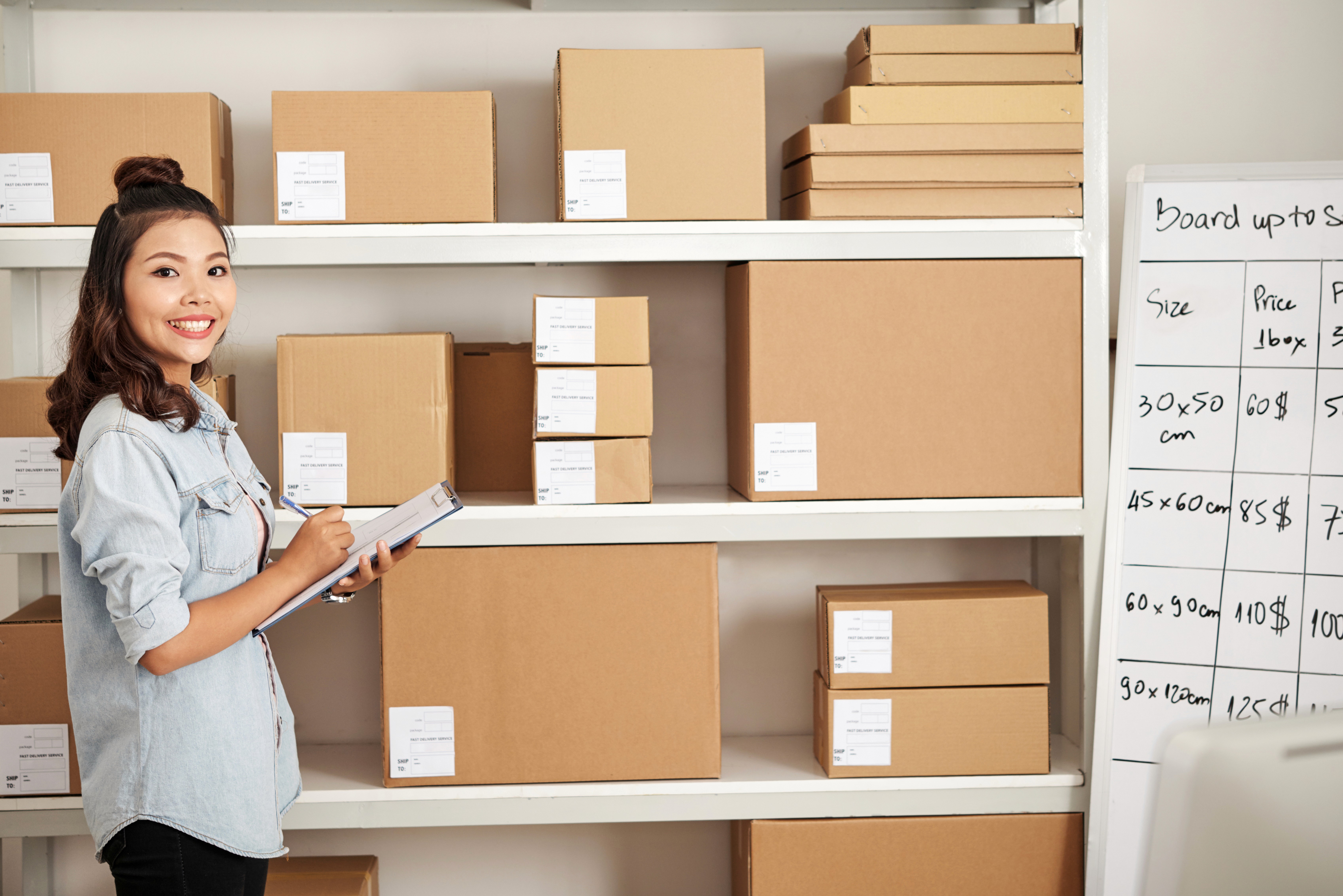 Smiling woman holding a clipboard stands in front of shelves filled with brown boxes of varying sizes. A whiteboard with product dimensions and prices is nearby.