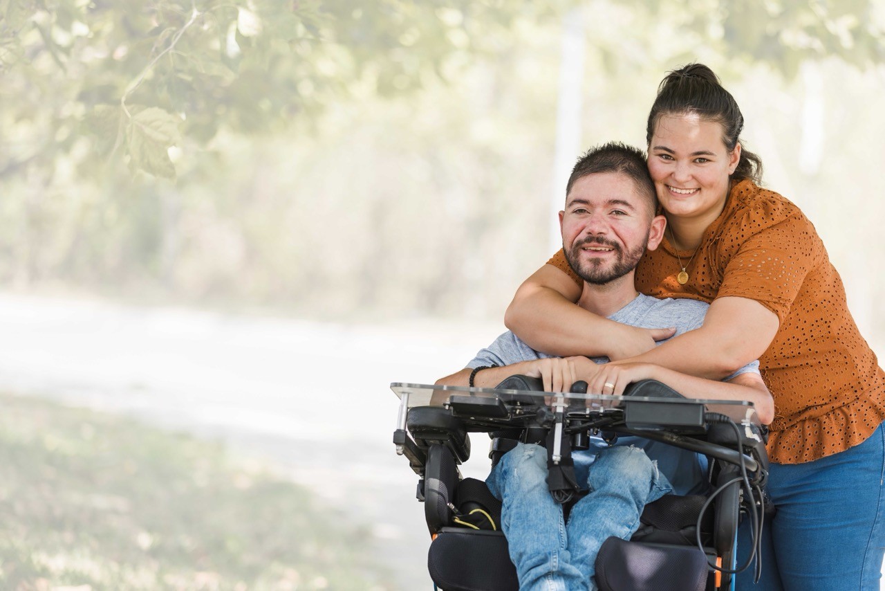 Background Image: A smiling woman hugs a man sitting in a motorized wheelchair outdoors on a sunny day, with green trees and a path in the background. Both appear happy and comfortable together.