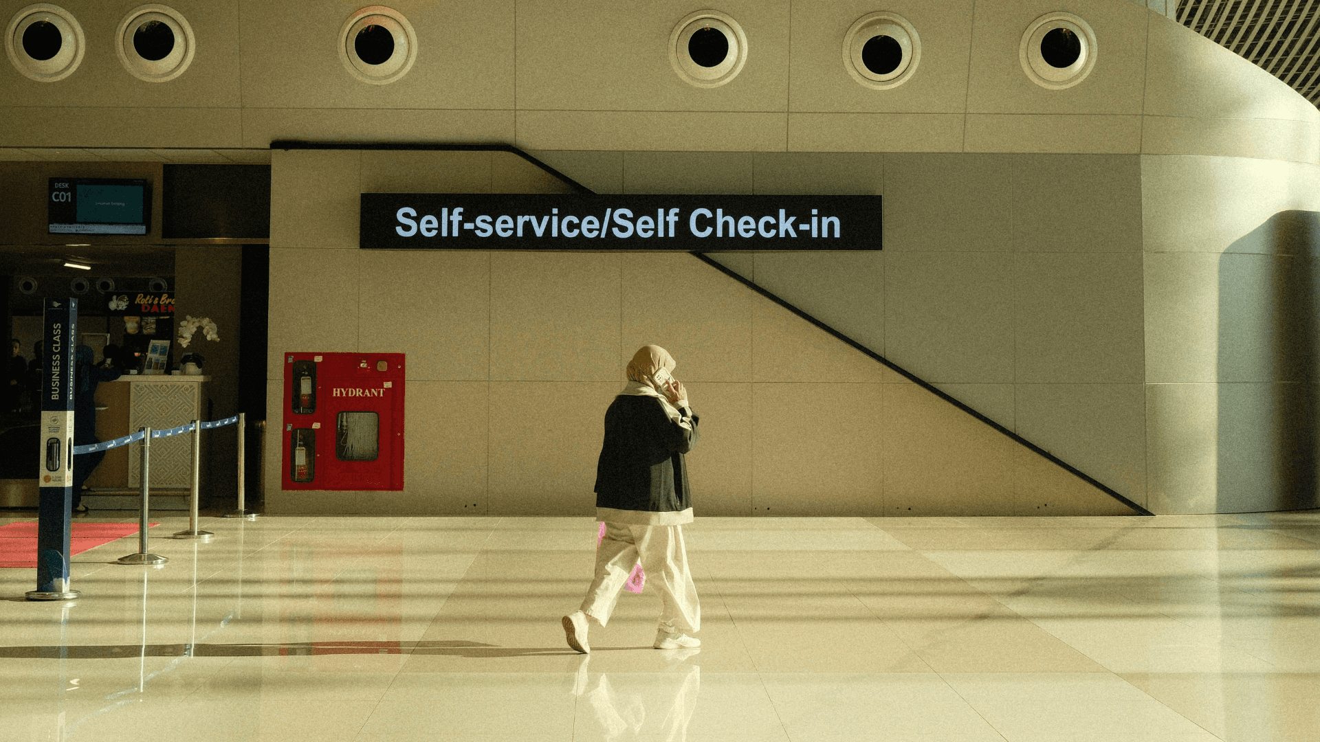A woman walks through an empty airport, the words 'self-service/self check-in' on a sign behind her
