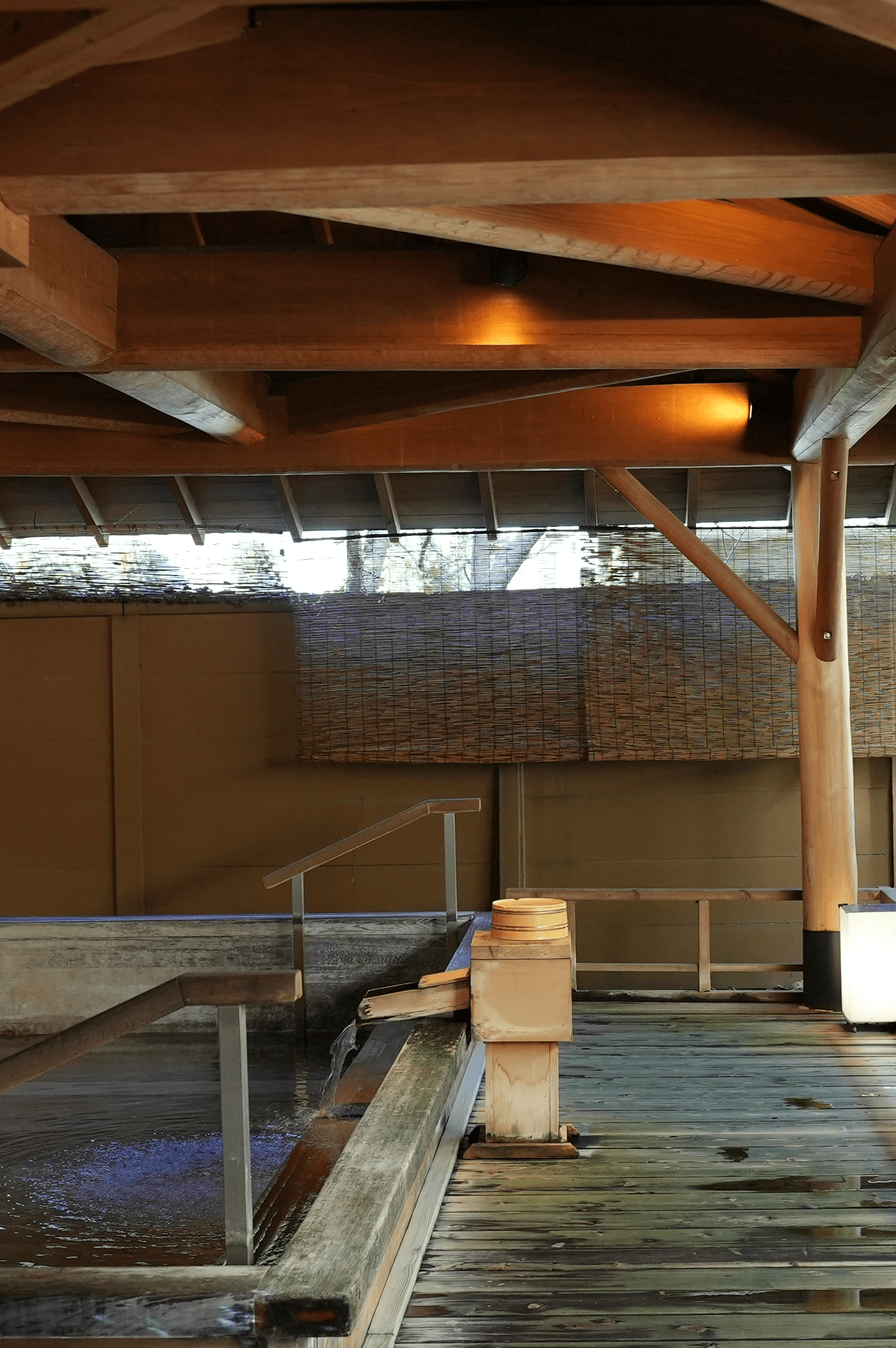 A serene outdoor Japanese hot spring (onsen) with a rock-lined pool of water, set under a traditional wooden pavilion with exposed beams. A small wooden spout pours water into the pool, which is surrounded by lush green plants and a stone wall.