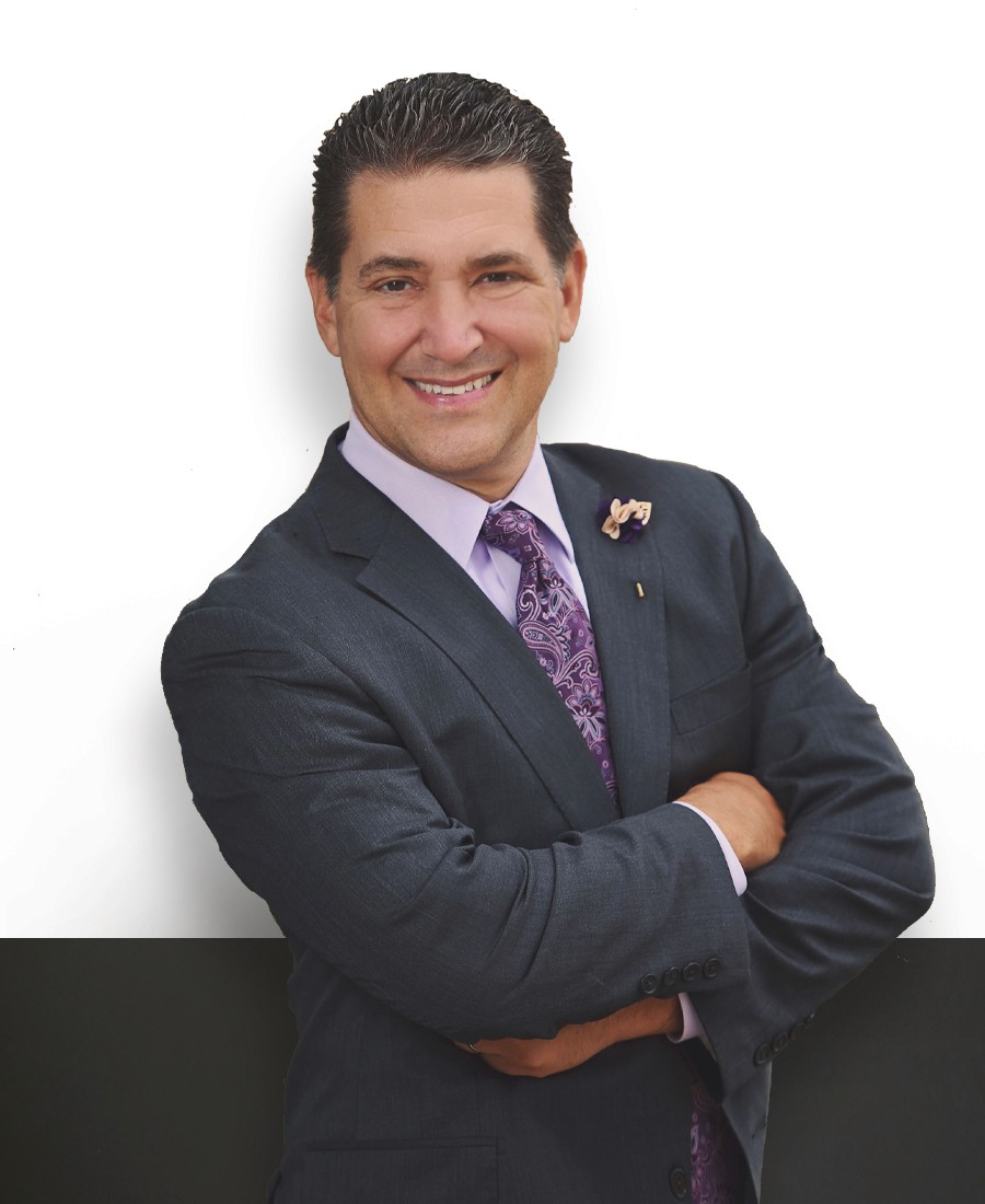 Professional headshot male with tan skin and black short hair wearing a dark grey suit and purple collared shirt with a purple tie and yellow  floral lapel pin, smiling confidently with arms crossed.