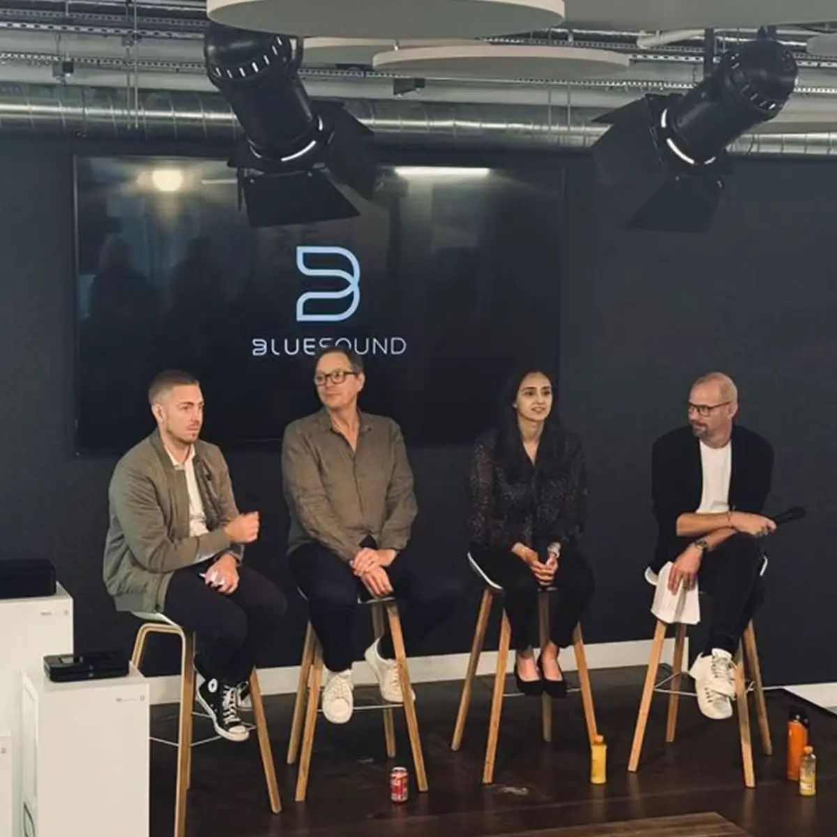 Panel discussion on a small stage with five speakers seated in front of a screen showing the Bluesound logo.