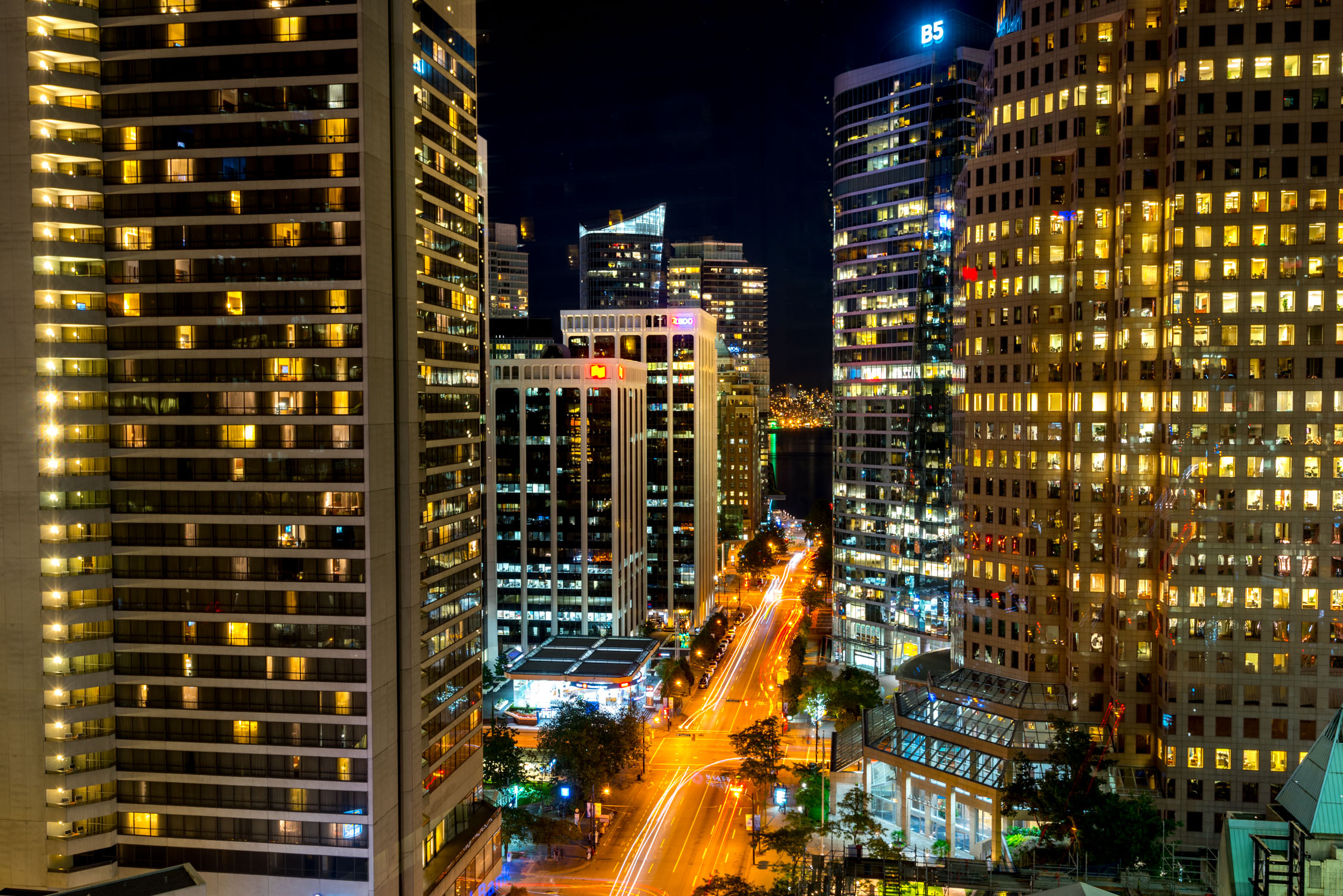Night view of illuminated skyscrapers and city streets with long-exposure light trails from traffic in a dense urban center.