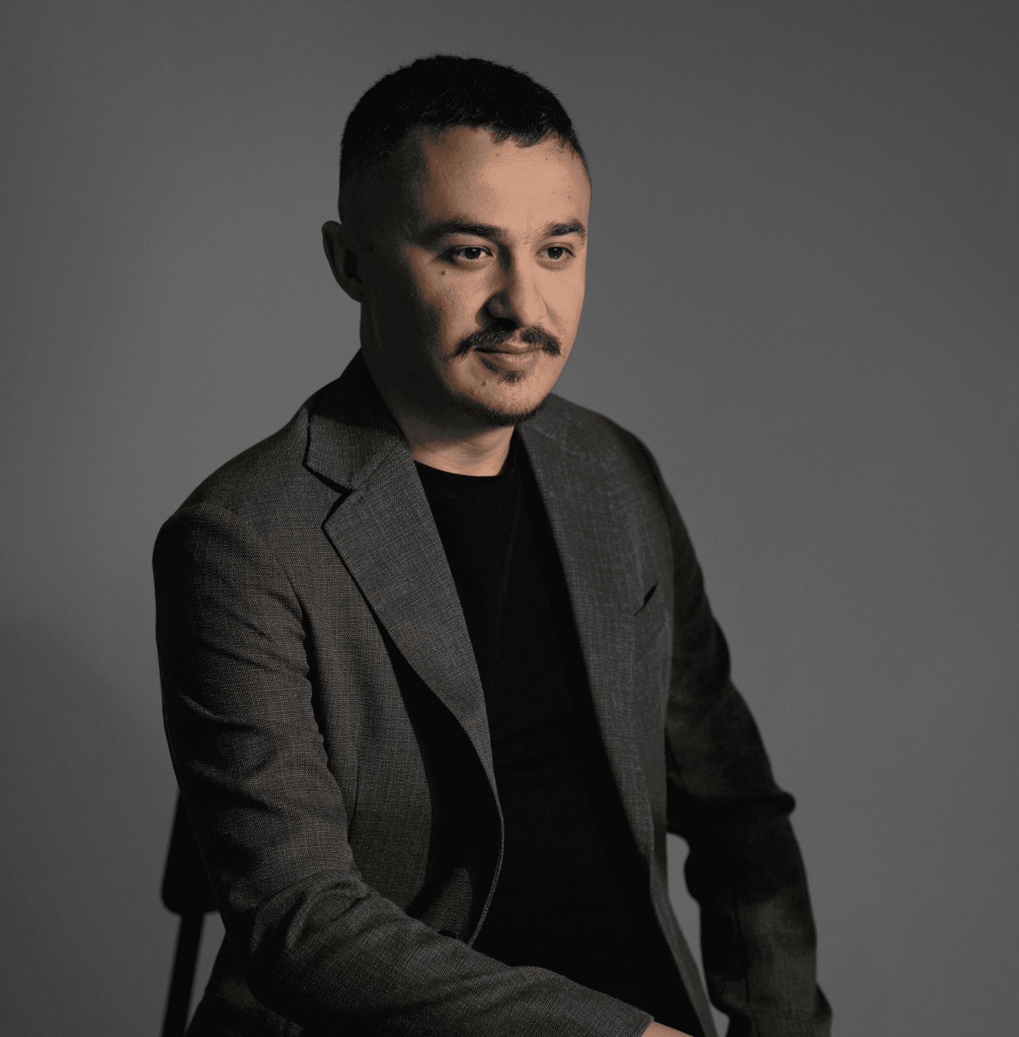 A person with short hair and a mustache, wearing a dark blazer over a black shirt, sits against a simple, dark background, conveying a professional and thoughtful demeanor.