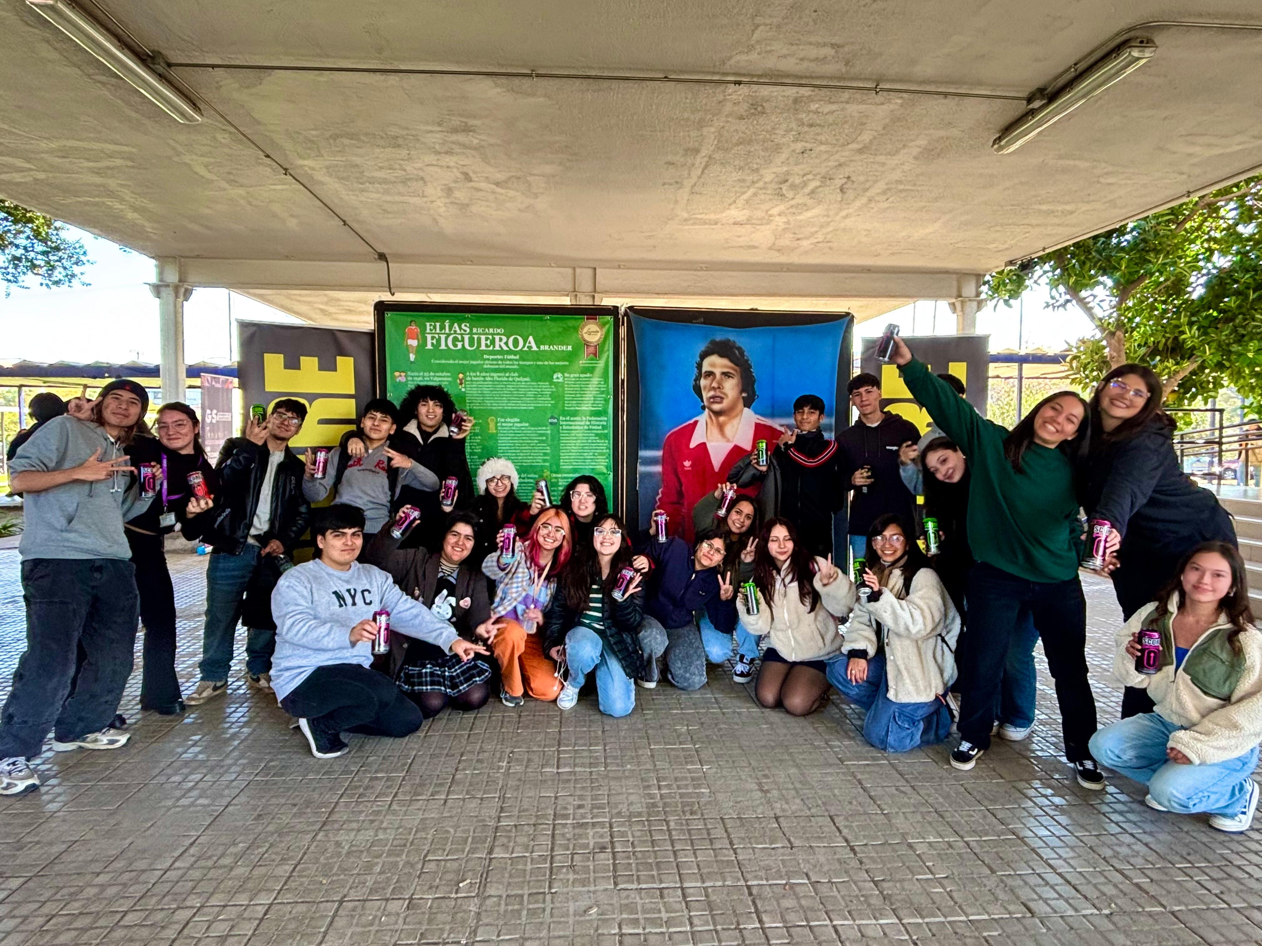 Grupo de personas con latas de Score Energy Drink frente a la pintura de Eliseo Salazar en la exposición Leyendas del Deporte USACH.