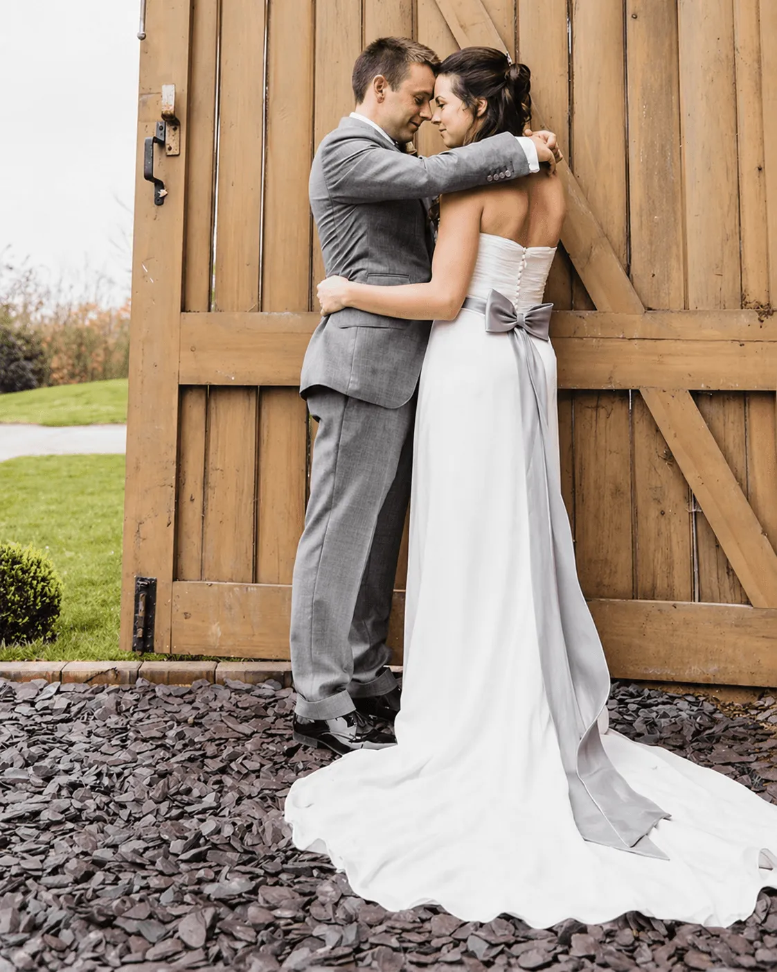 A smiling groom in a brown suit is embraced by his bride in a white,  lace wedding dress