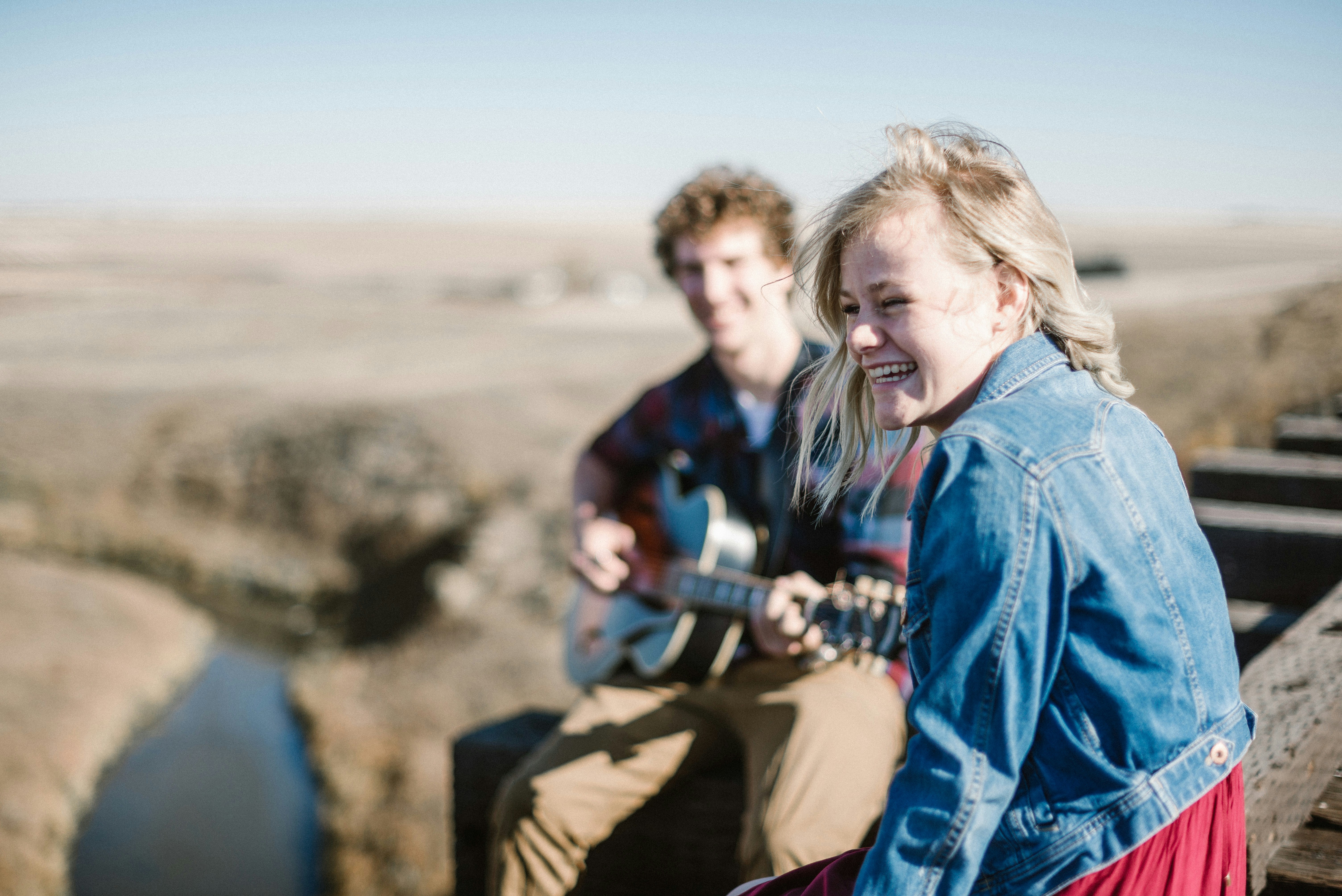 A woman wearing a denim jacket looks to her left. Behind her, a man plays the guitar. The background shows a distant valley and river.