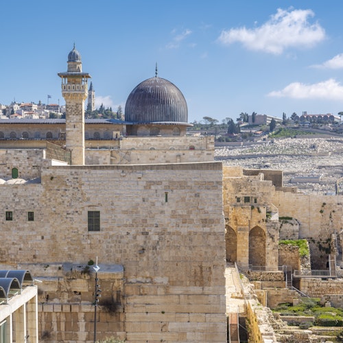 Historic stone building complex with a large dome and a minaret under a bright blue sky with scattered clouds.