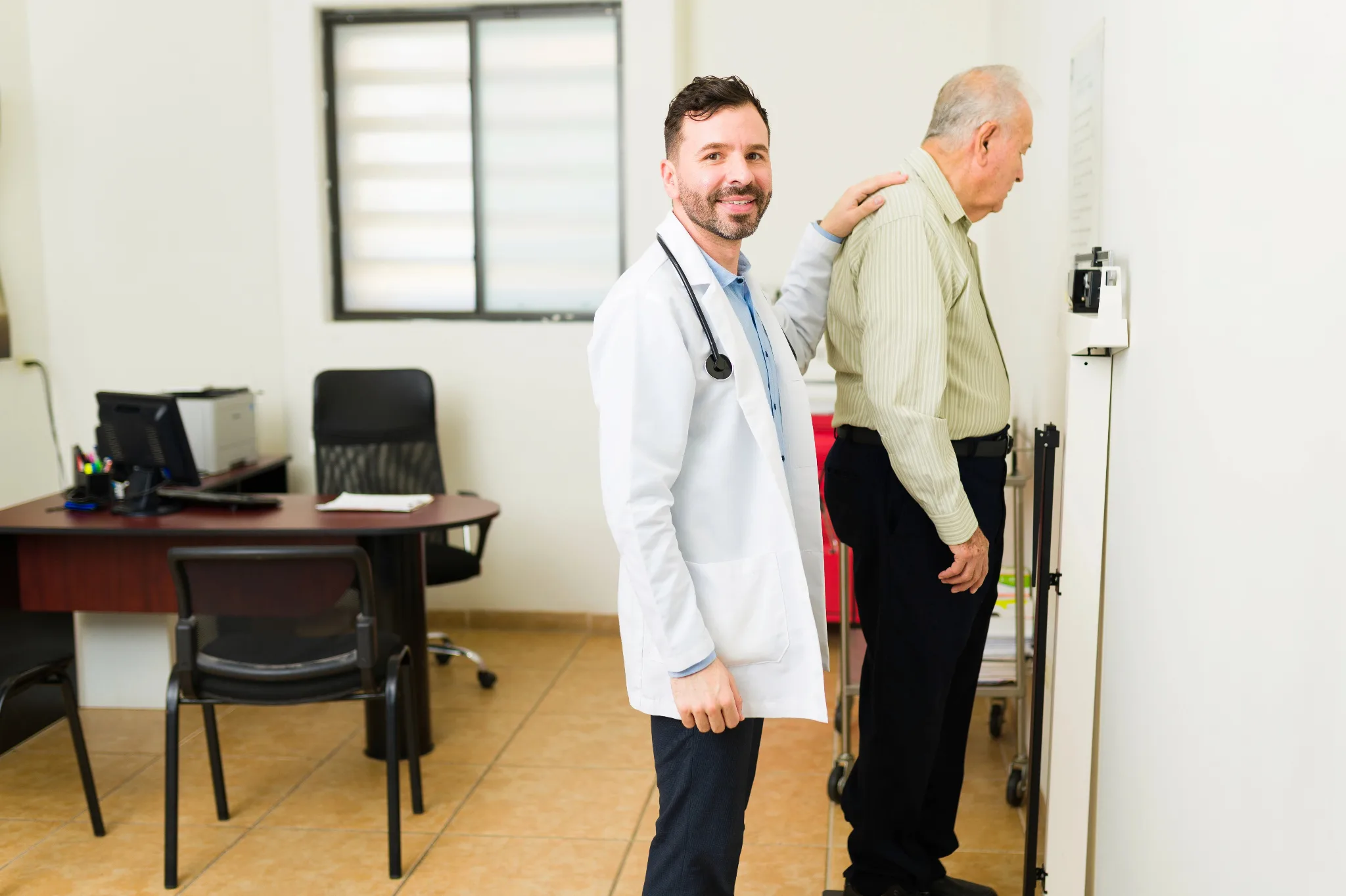 A male doctor in a white coat with a stethoscope examines an elderly patient's posture and shoulder mobility in a clinical examination room.