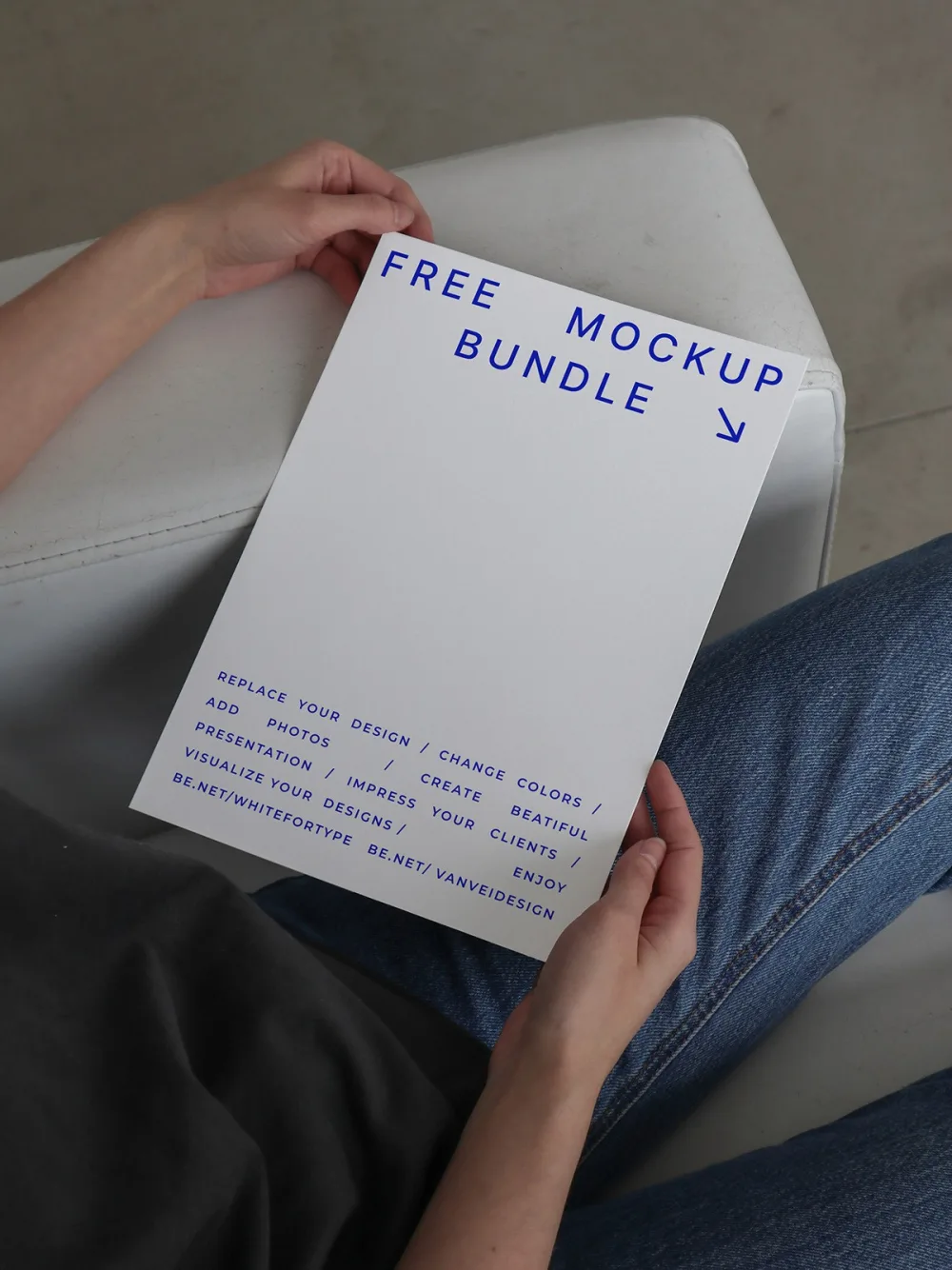 A person holding a white A4 paper mockup while seated on a white chair, wearing blue jeans and a dark gray shirt. The setting is minimalistic, with soft natural lighting enhancing the neutral tones.