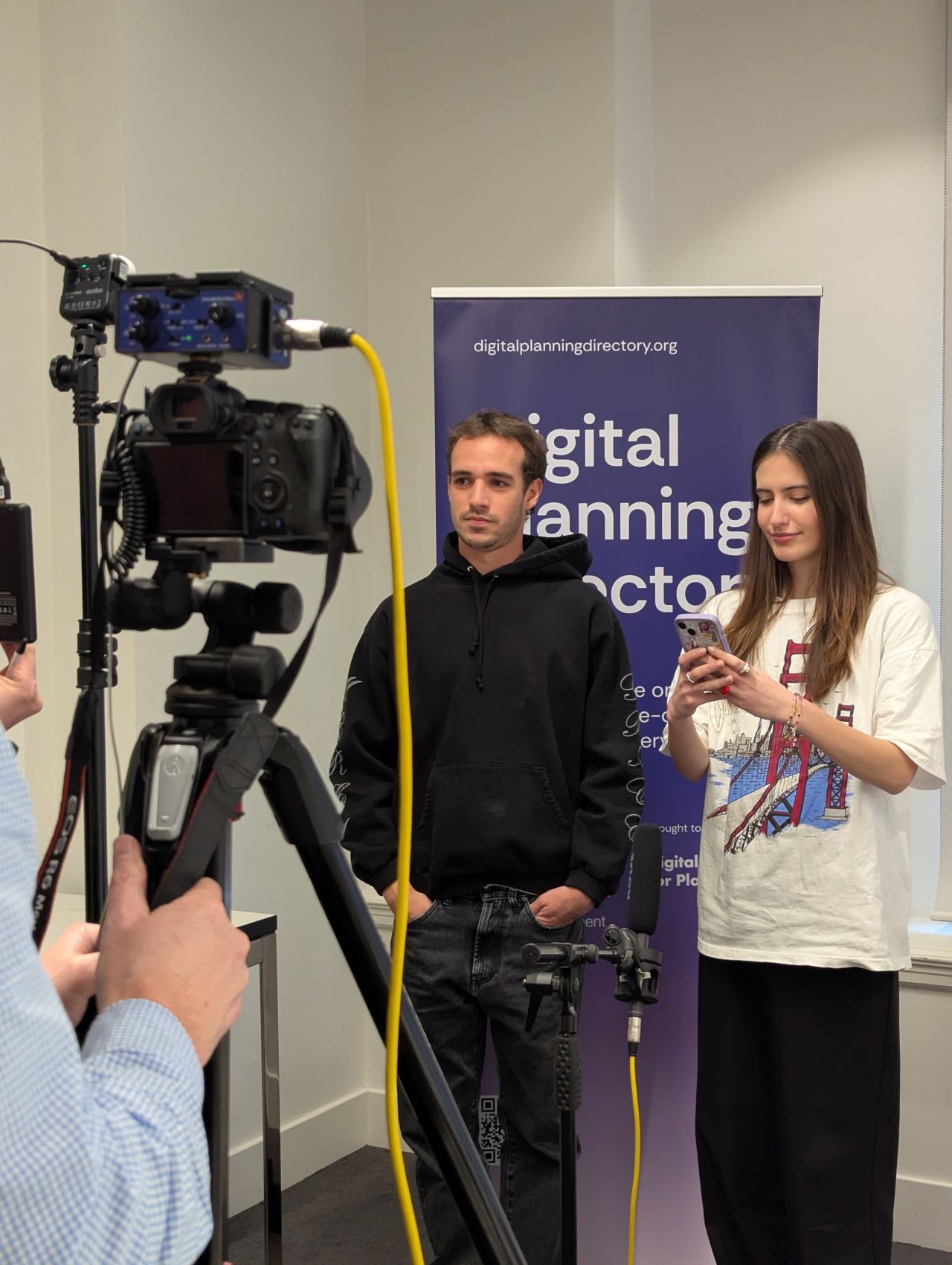 A young man in a black hoodie being filmed at the Digital Planning Directory Launch, with a woman beside him holding a phone and a professional camera recording.