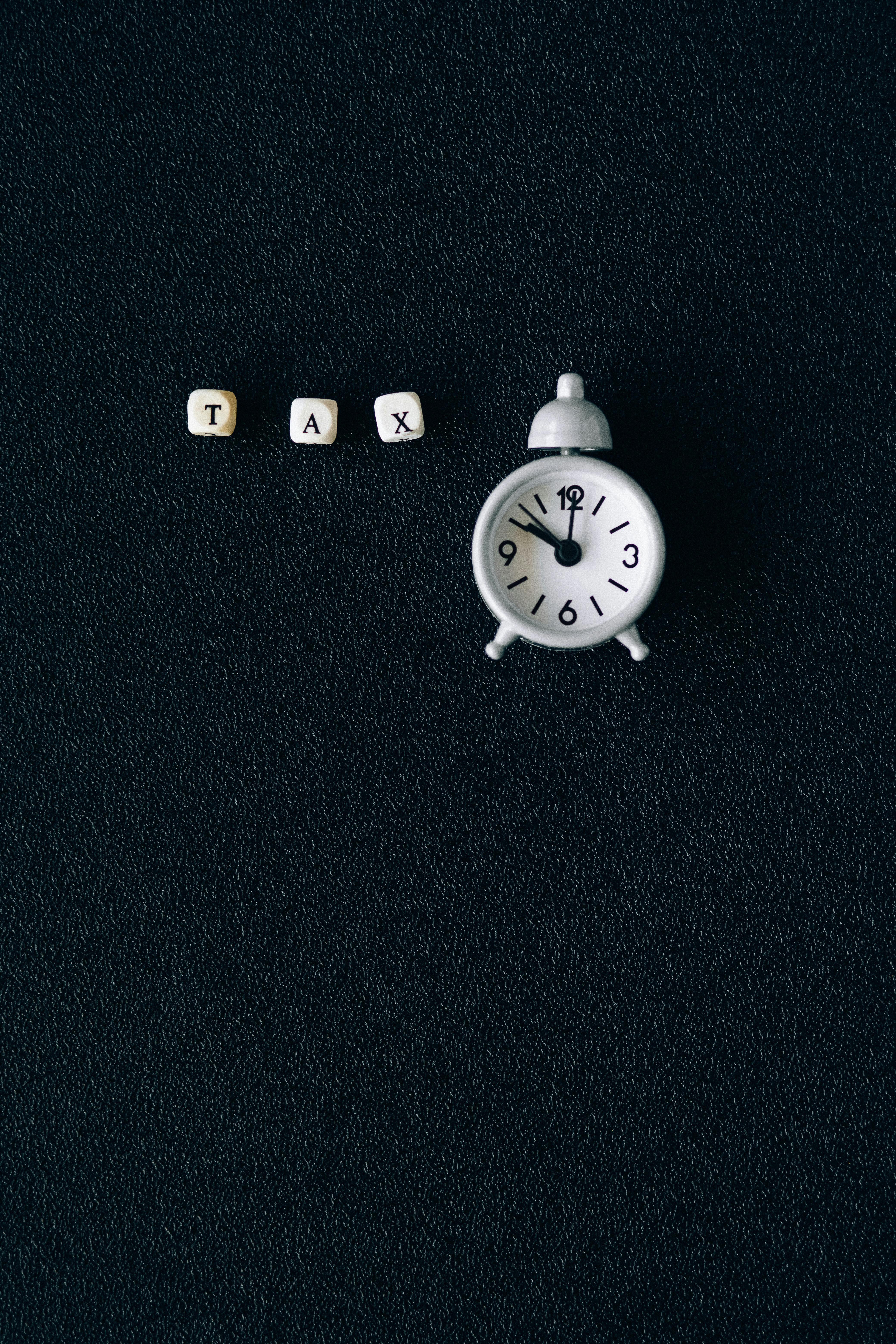 Small white letter cubes spelling the word "TAX" next to a small, white vintage-style alarm clock.